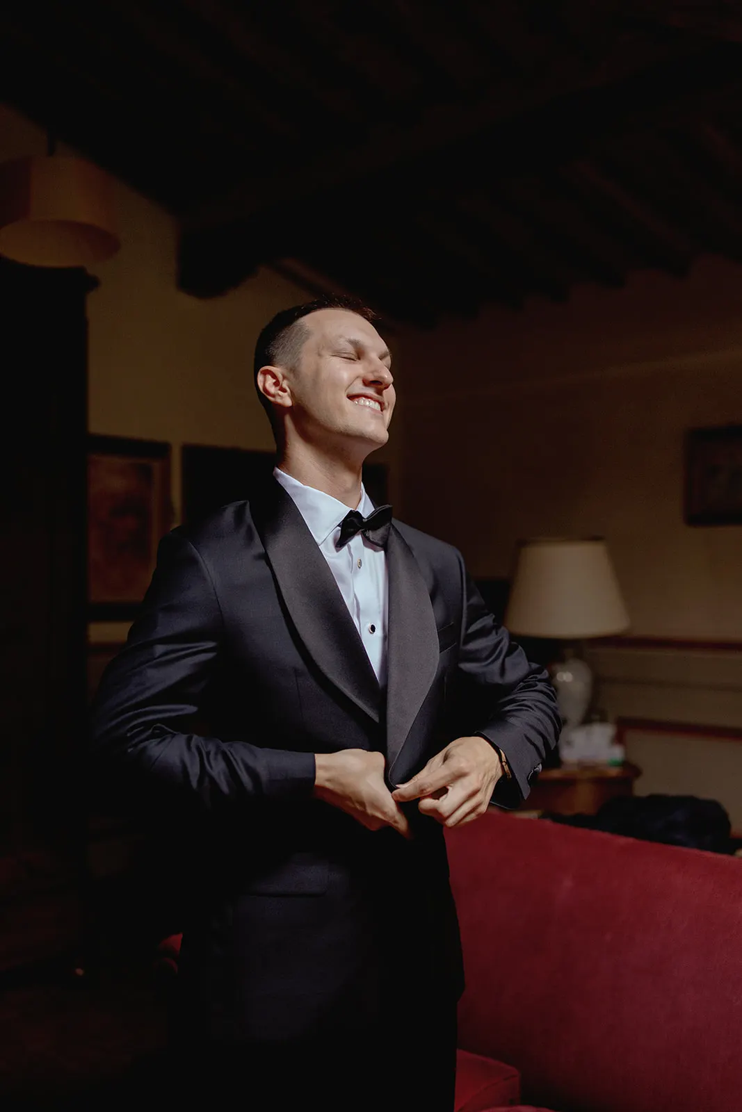 A man in a dark tuxedo with a bow tie stands indoors, smiling while buttoning his jacket. The room has warm lighting, a red couch, a white lamp, and framed art on the walls. The ceiling features exposed wooden beams. Tuscan Wedding Photographer