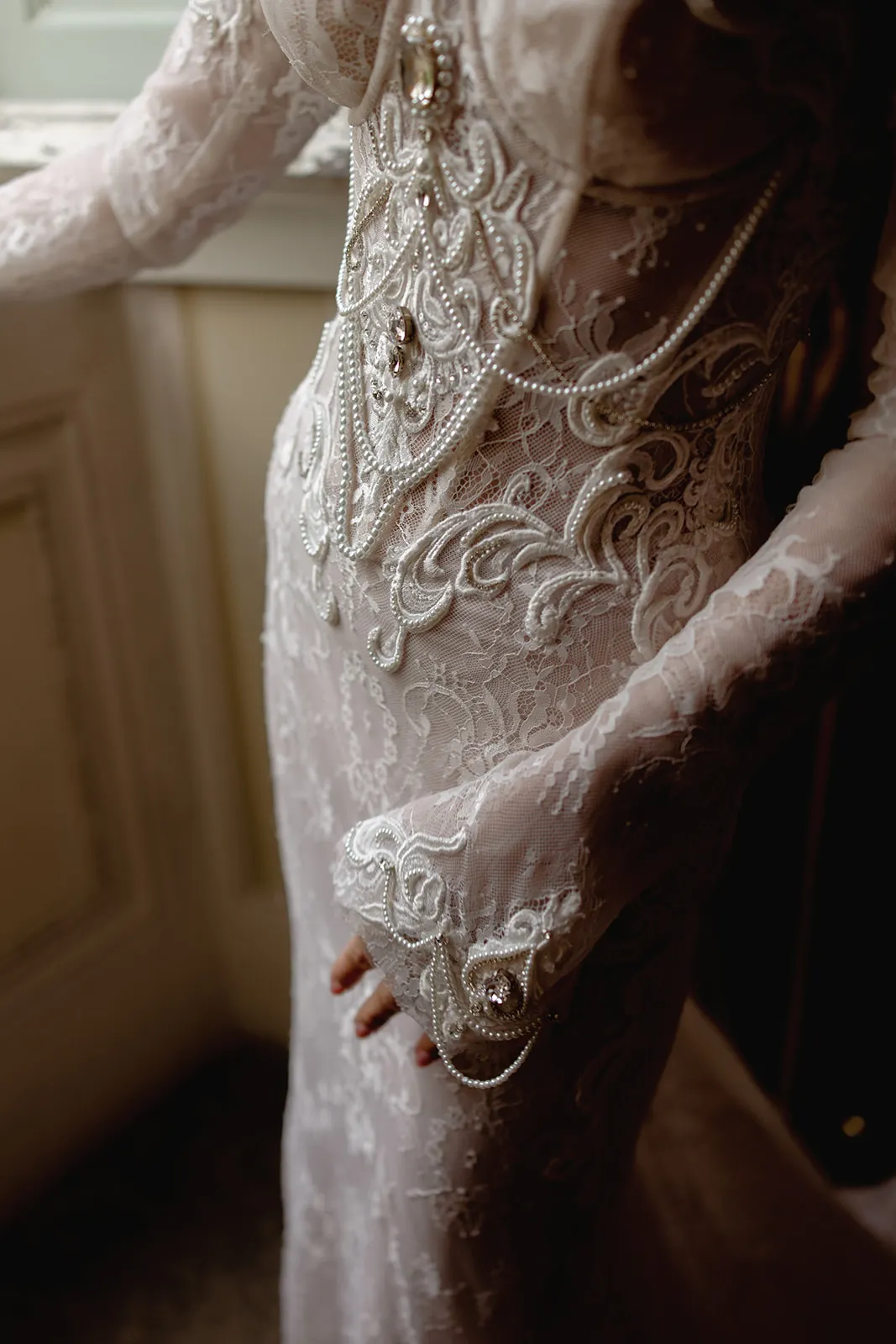A close-up of a person in a fitted, sheer lace wedding dress with intricate embroidery, pearl details, and bead embellishments. The sleeves are long and decorated with lace and pearls. The figure stands by a window, softly lit by natural light. Tuscan Wedding Photographer