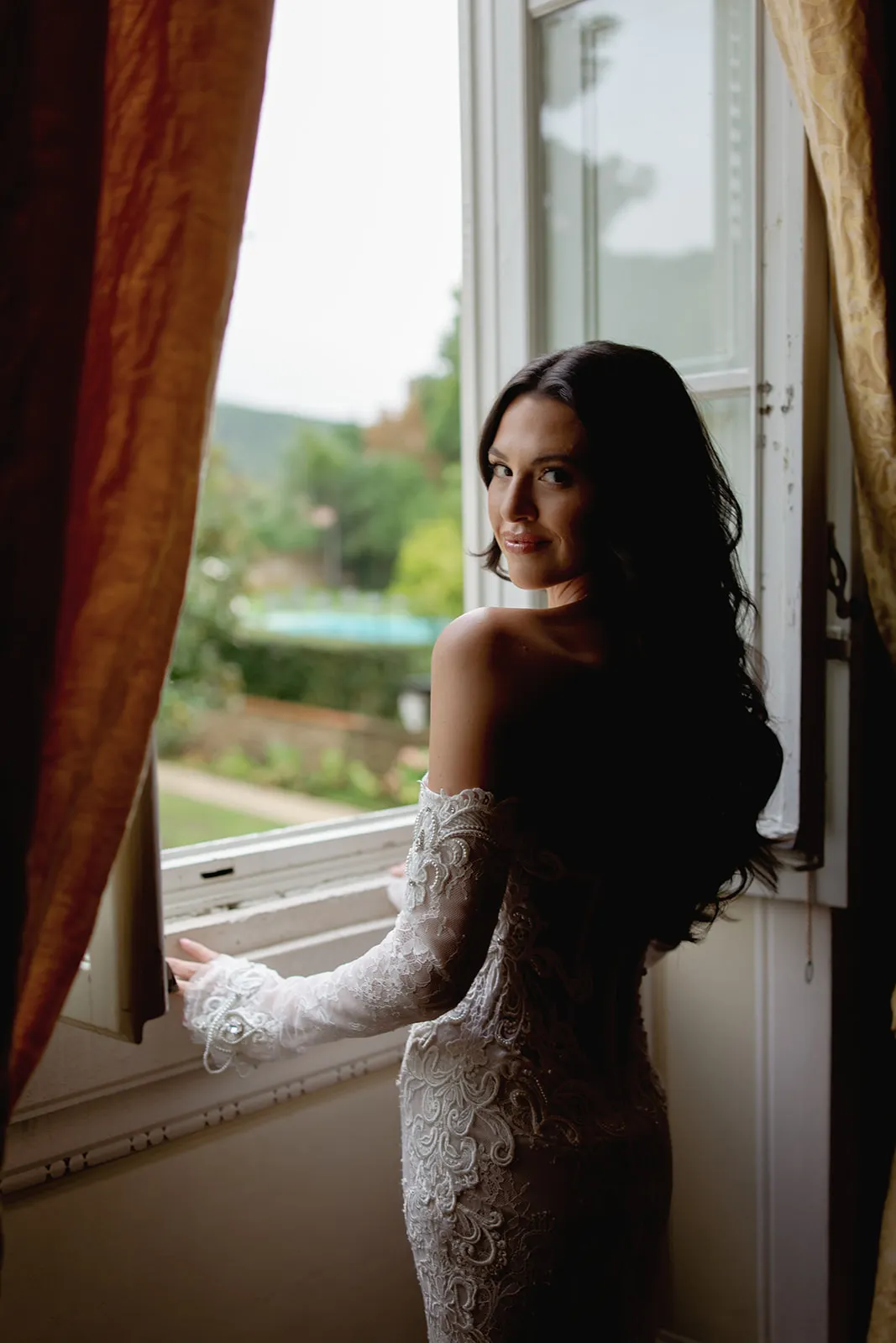 A woman with long dark hair in an off-shoulder, lace white gown stands by an open window, looking back and smiling. Lush green scenery is visible outside. Warm, gold curtains frame the window, and soft natural light illuminates the scene. Tuscan Wedding Photographer