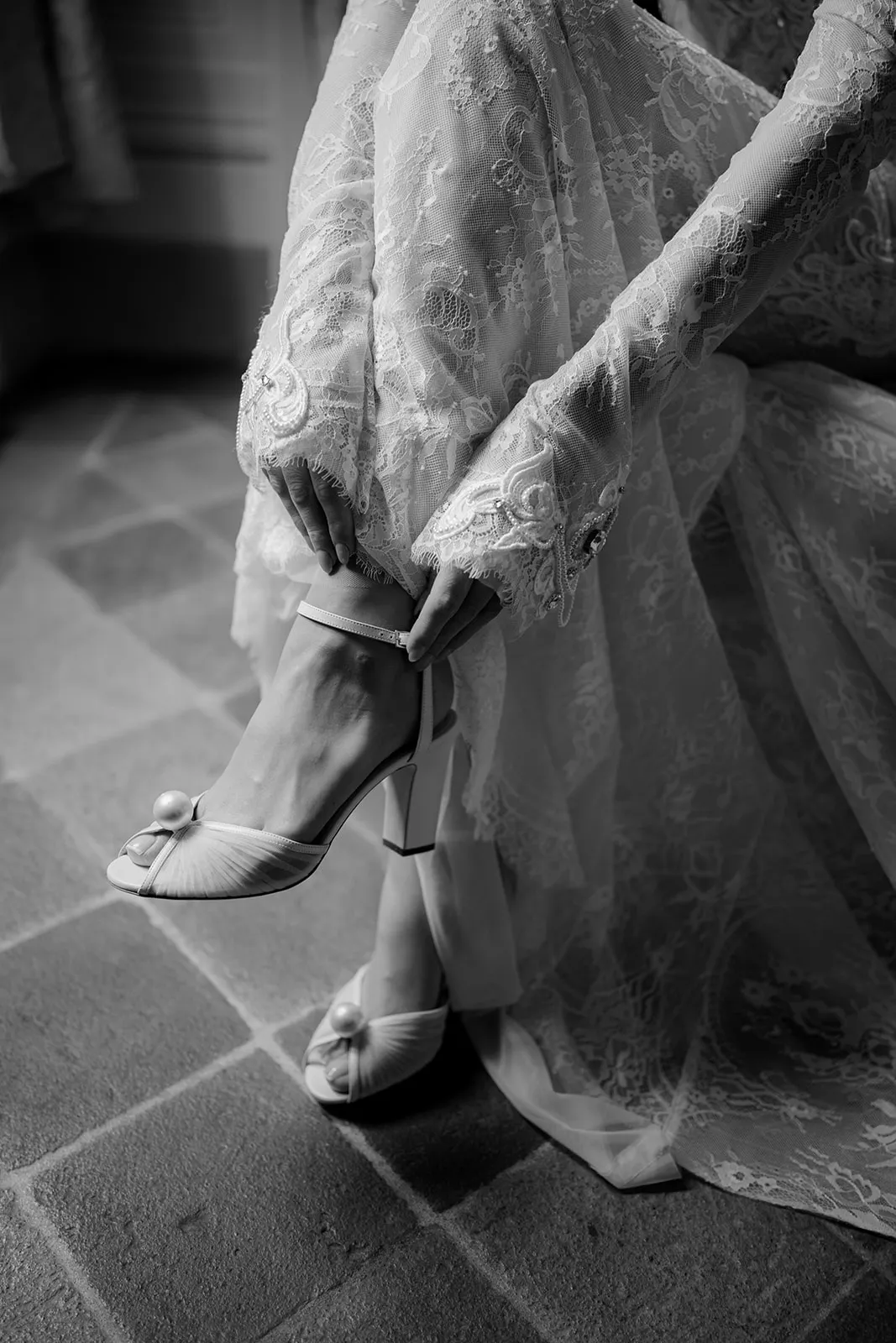 A person in a long, lace wedding dress adjusts a high-heeled, open-toed shoe with a decorative pearl. The image is in black and white, showing delicate lace sleeves and a tiled floor beneath their feet. Tuscan Wedding Photographer