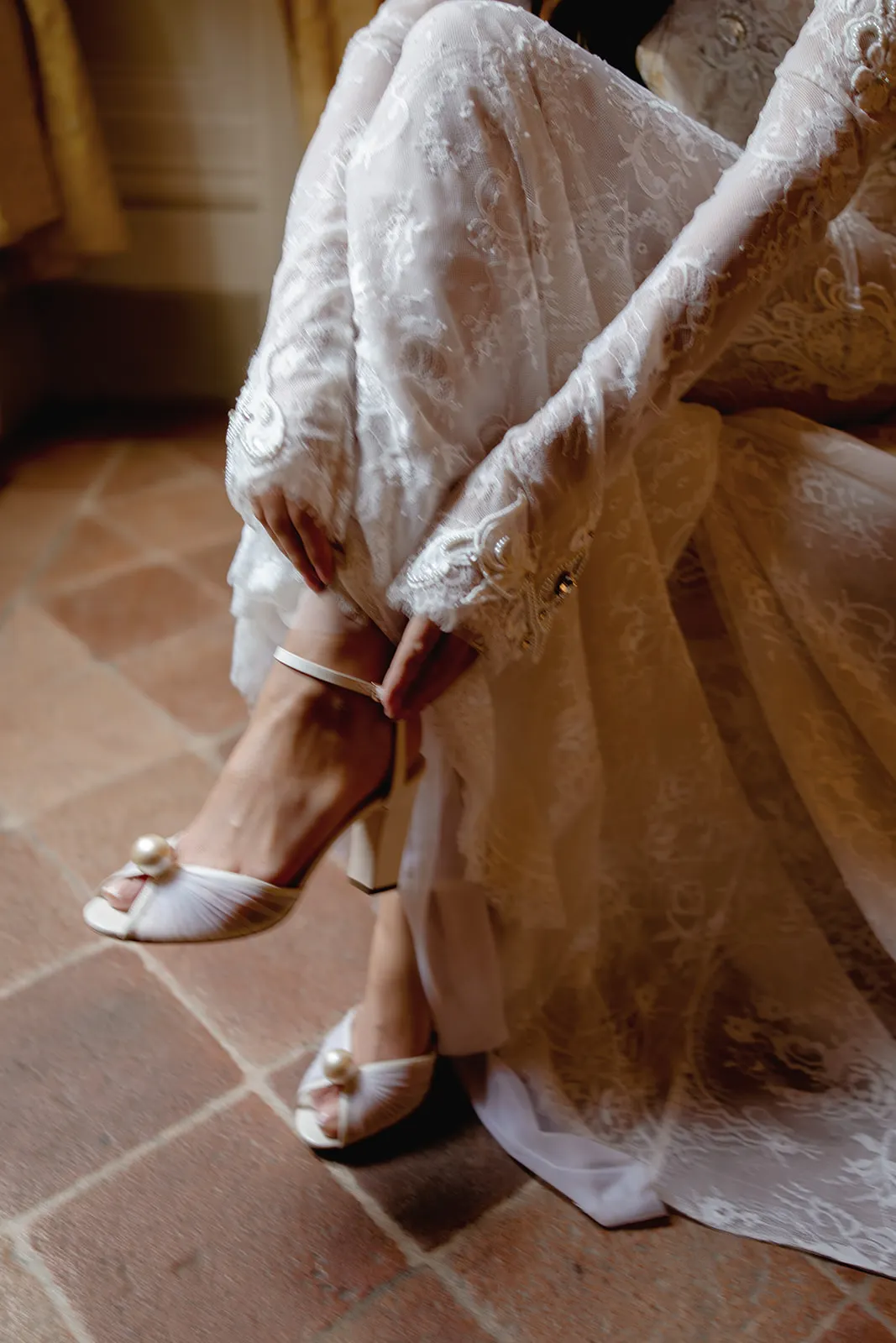 A person in a delicate, lace long-sleeve white dress sits on a tiled floor, adjusting a high-heeled sandal with ankle straps and pearl embellishments. The scene is softly lit, evoking an elegant, serene, and possibly bridal moment. Tuscan Wedding Photographer