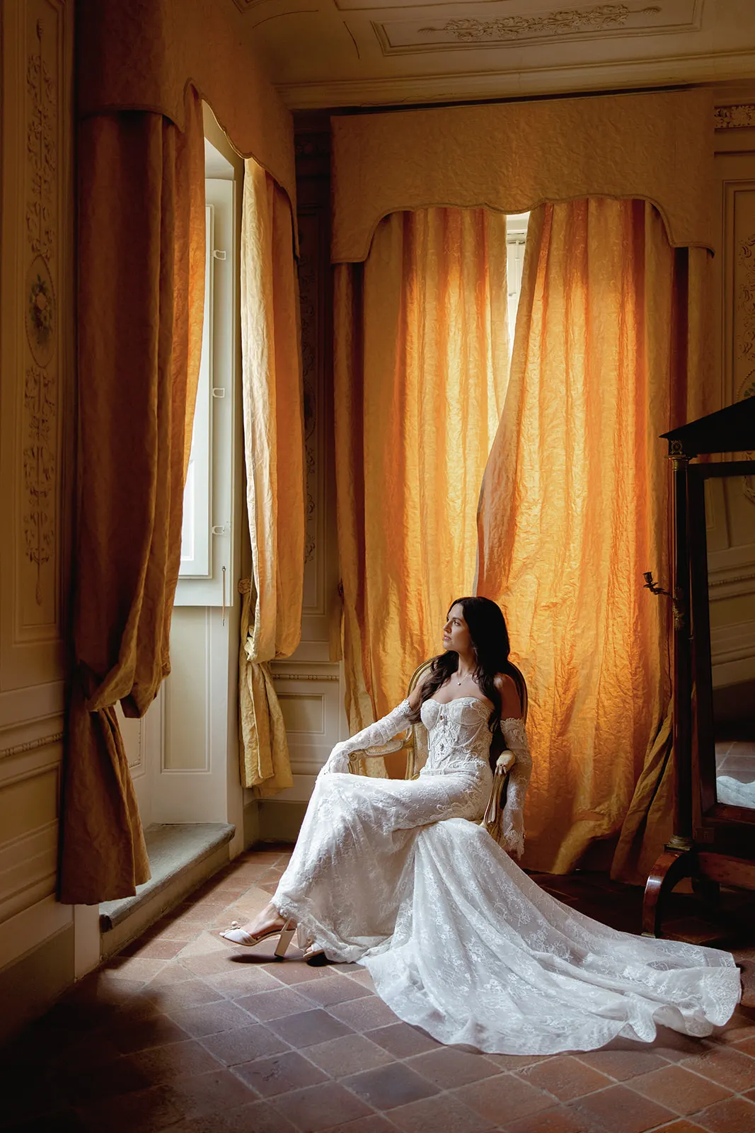 A woman in a long, white lace wedding gown with a train sits on an ornate chair, gazing out a tall window. Warm golden curtains and soft natural light fill the elegant, vintage-style room, creating a serene and dreamy atmosphere. Tuscan Wedding Photographer