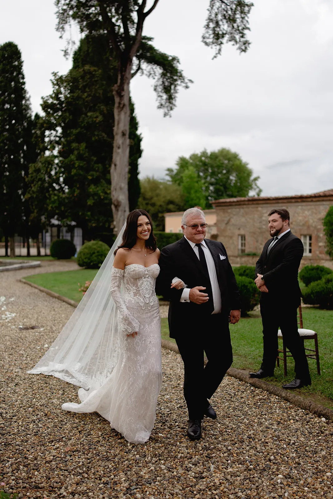 A bride in a white lace gown and long veil walks arm-in-arm with an older man in a black suit on a gravel path outdoors. Both are smiling. Tall trees, green grass, and stone buildings are visible in the background. A man in black stands nearby, watching. Tuscan Wedding Photographer