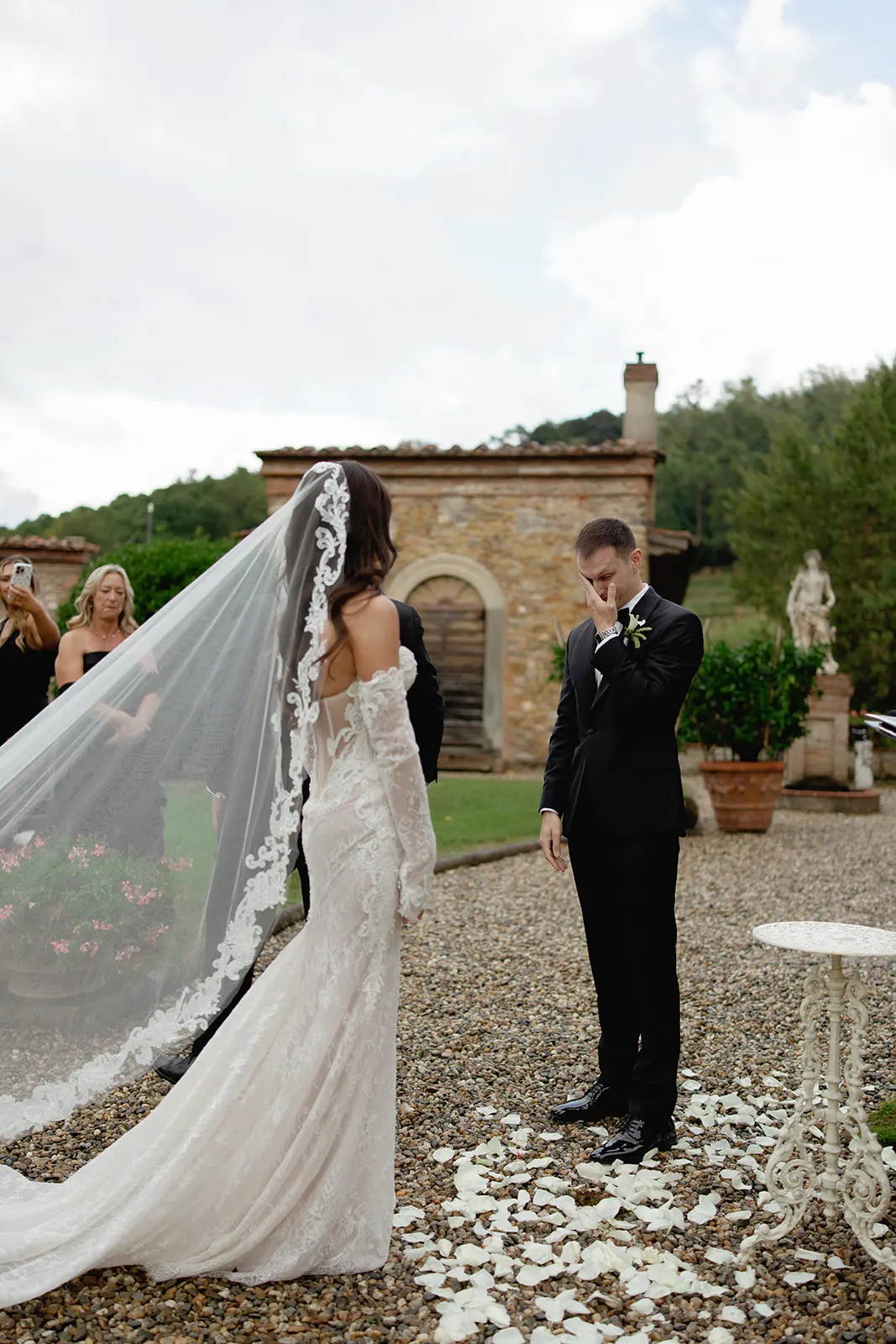A bride in a white lace gown with a long, floral veil stands facing a groom in a black suit who wipes his eyes, appearing emotional. They are outdoors on a gravel path with scattered flower petals, surrounded by greenery and stone buildings. Tuscan Wedding Photographer