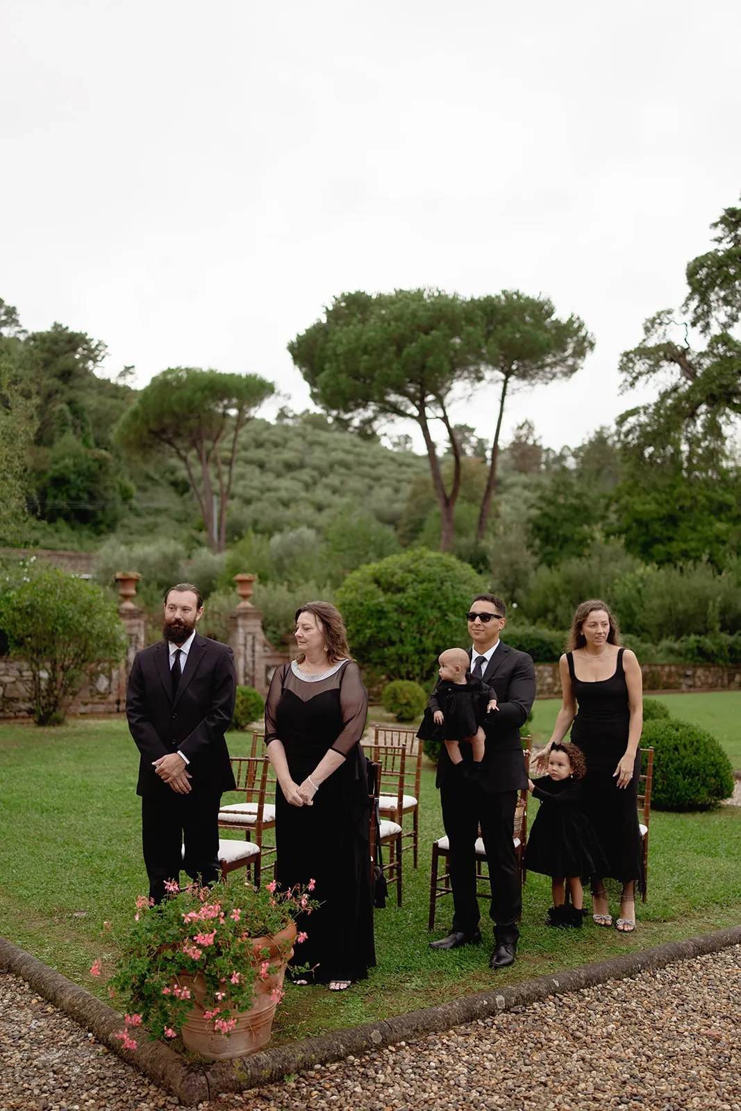Five people stand on grass at an outdoor event, all dressed in formal black attire. Two men and three women are present; one man holds a baby, and a woman holds a child’s hand. Trees and greenery surround the area; chairs and flowers are visible. Tuscan Wedding Photographer
