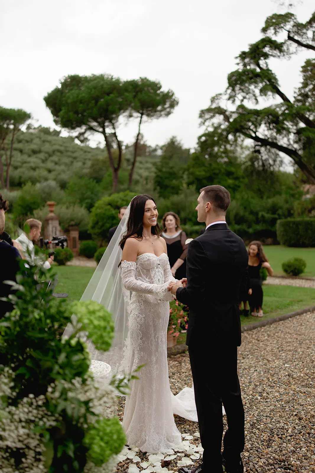 A bride in an off-shoulder lace gown and veil smiles at her groom, who wears a black suit, during an outdoor wedding ceremony. They stand on a gravel path, holding hands, with greenery and seated guests in the background on a cloudy day. Tuscan Wedding Photographer