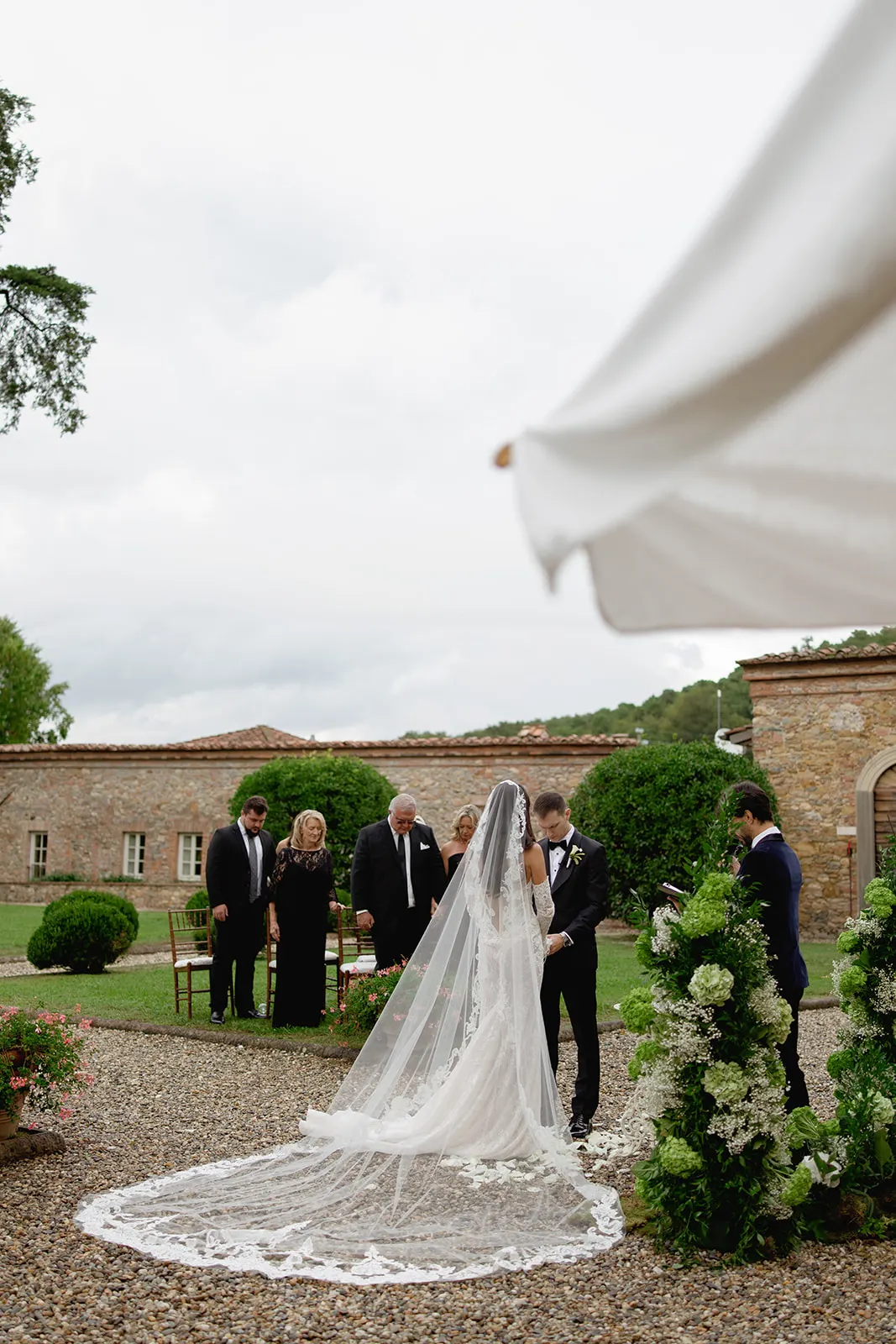 A bride in a long white gown with a lace train and veil stands with the groom at an outdoor wedding ceremony. They face each other near a flower arch; guests in formal attire sit and stand behind them amid stone buildings and greenery. Tuscan Wedding Photographer