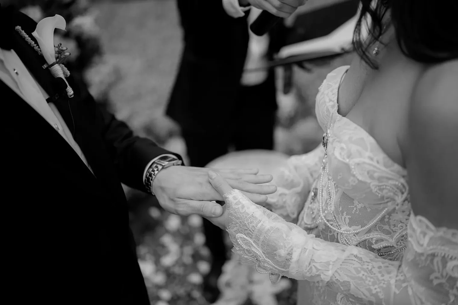 A black-and-white close-up photo of a couple at their wedding ceremony. The bride in a lacy off-shoulder gown touches the grooms hand. The groom wears a suit with a calla lily boutonniere. An officiant stands blurred in the background, holding a microphone. Tuscan Wedding Photographer