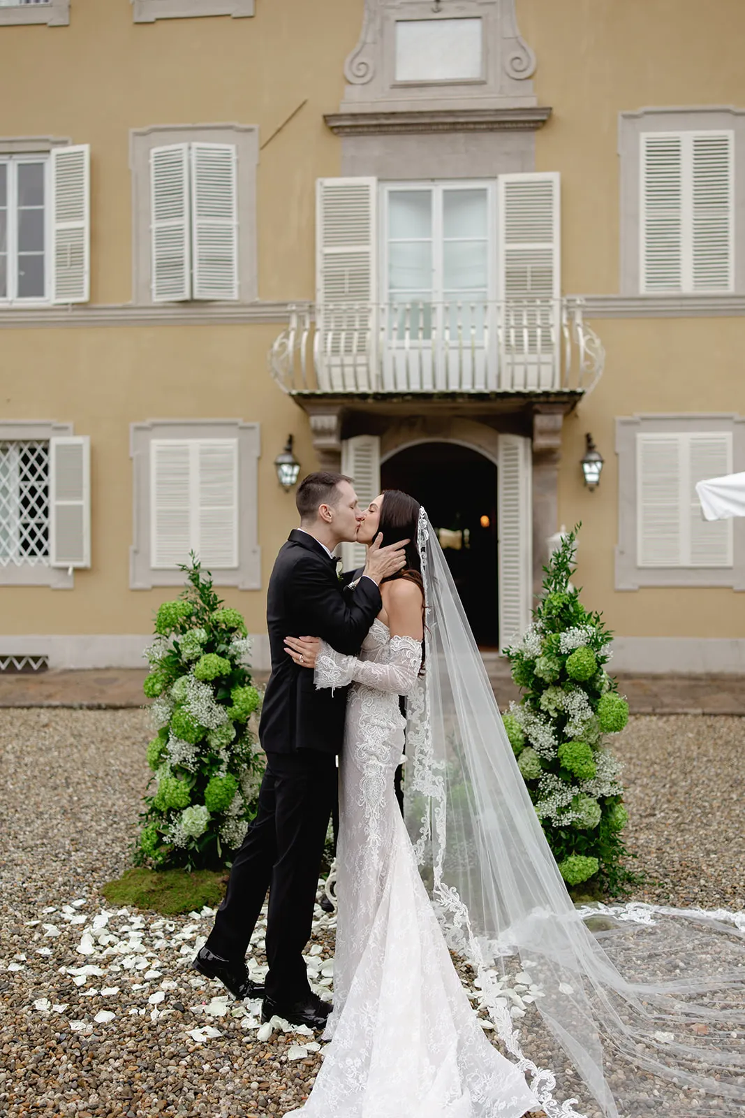 A couple in wedding attire kisses outdoors in front of an elegant building with white shutters. The bride wears a long, white gown with a veil, and the groom wears a black suit. Green floral arrangements and white petals decorate the stone courtyard. Tuscan Wedding Photographer