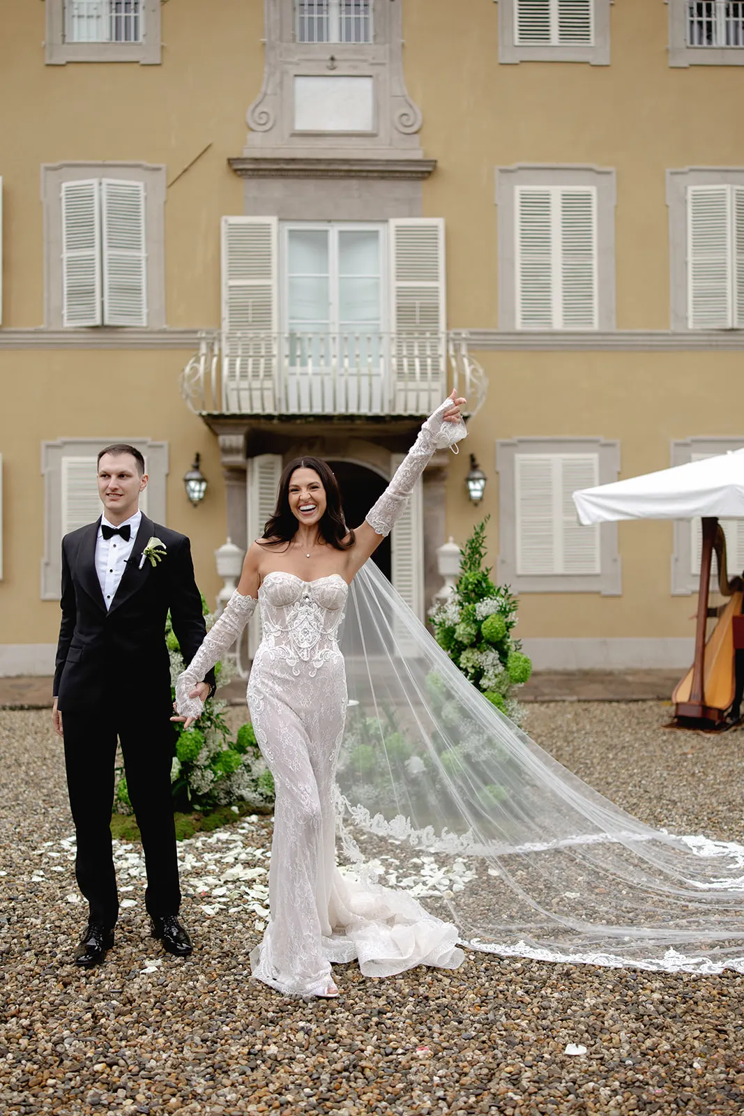 A bride in a lacy, strapless white gown with a long veil and gloves smiles and raises one arm, holding hands with a groom in a black tuxedo. They stand on gravel outside a tan building with white shutters and flower petals scattered on the ground. Tuscan Wedding Photographer