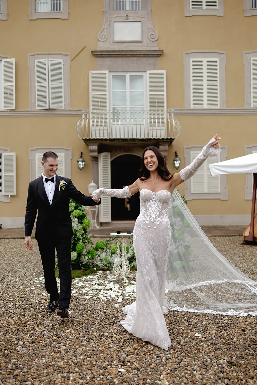 A joyful bride in a lacy, off-shoulder white gown with a long veil holds hands with a smiling groom in a black tuxedo as they walk outside a grand building with cream walls and white shutters. Flowers and petals decorate the ground behind them. Tuscan Wedding Photographer