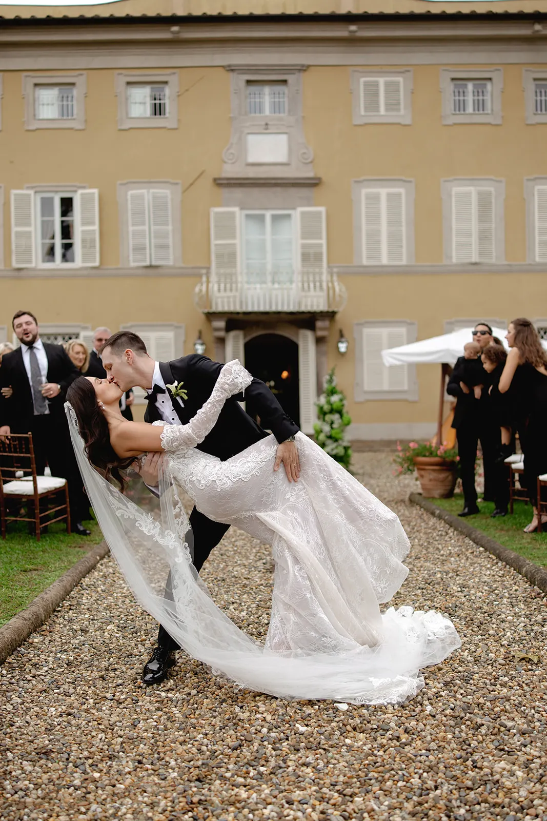 A bride and groom share a dramatic kiss as the groom dips the bride on a gravel path. The bride wears a lacy white gown and veil; guests watch and smile. The backdrop is a large, tan villa with shuttered windows and greenery. Tuscan Wedding Photographer