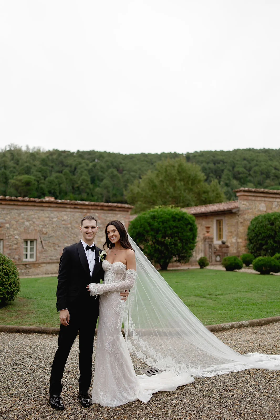 A bride in a strapless white gown with a long veil and a groom in a black tuxedo stand smiling together outdoors on a gravel path, with green grass, manicured bushes, and a rustic stone building surrounded by trees in the background. Tuscan Wedding Photographer