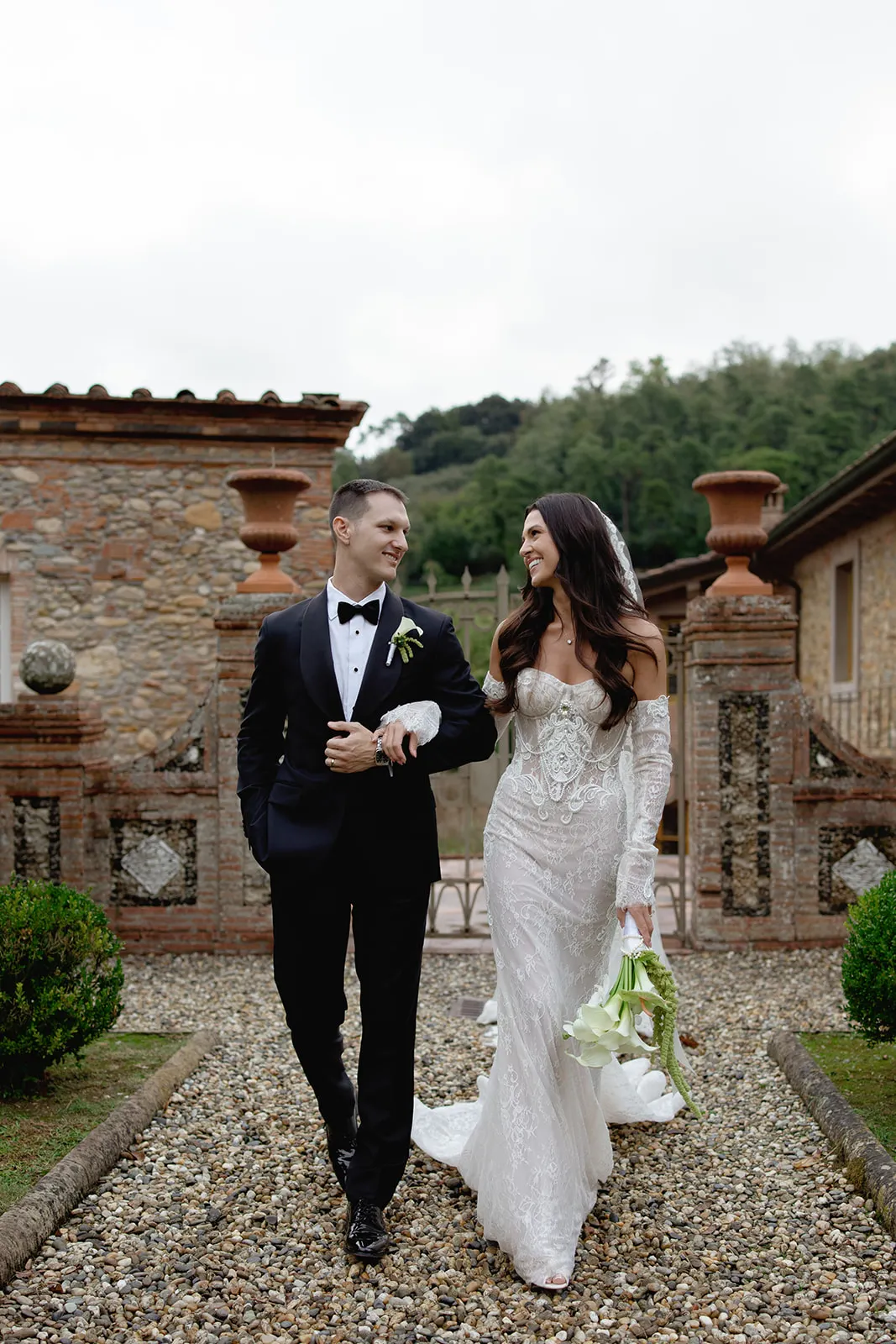A bride in a white, off-the-shoulder lace gown and veil holds calla lilies, walking arm-in-arm with a groom in a black tuxedo. They smile at each other on a pebble path, flanked by greenery and stone buildings, with trees in the background. Tuscan Wedding Photographer