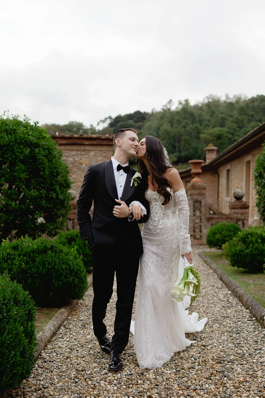 A bride in an off-shoulder lace gown and a groom in a black tuxedo walk arm in arm on a gravel path, surrounded by manicured bushes and rustic buildings. The bride kisses the grooms cheek while holding a bouquet of white flowers. Tuscan Wedding Photographer