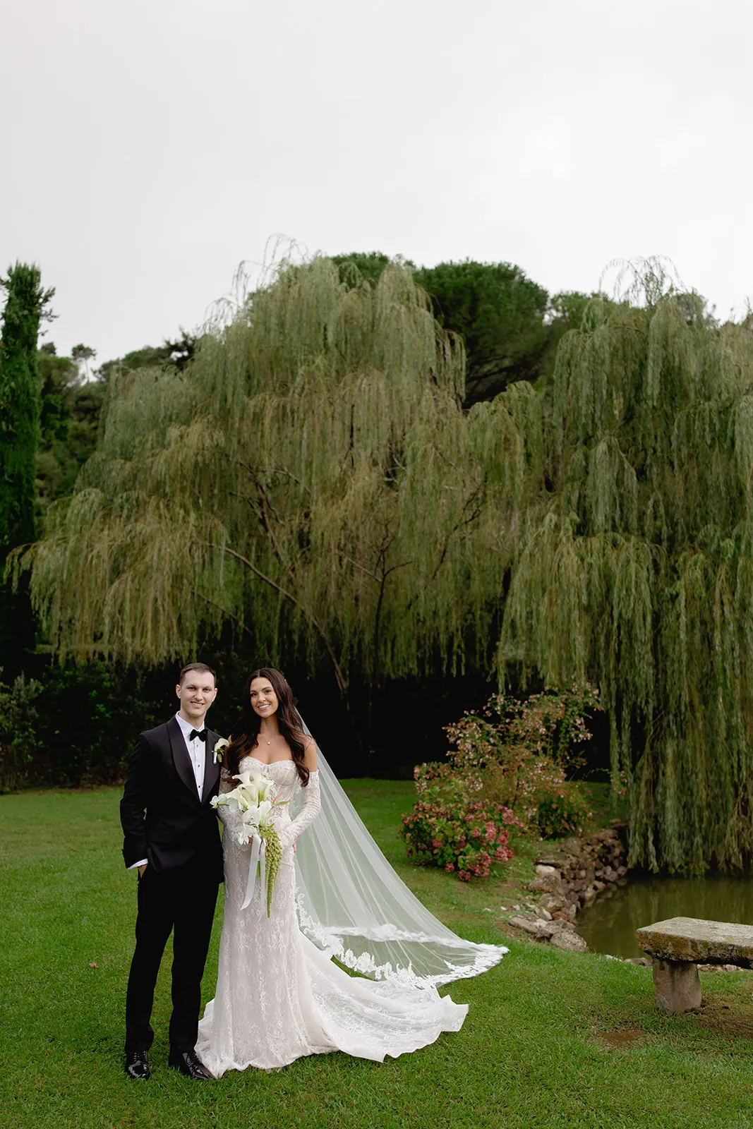 A bride in a long white lace gown with a flowing veil stands beside a groom in a black suit and bow tie. They smile outdoors on green grass, with a pond, blooming bushes, and tall willow trees in the background under an overcast sky. Tuscan Wedding Photographer