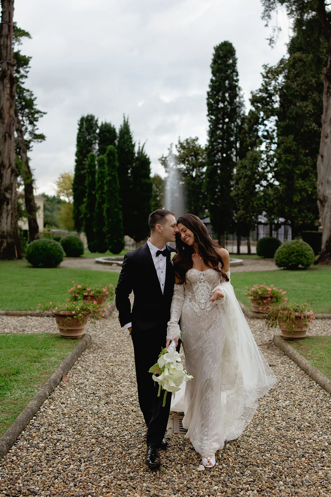 A bride and groom walk arm-in-arm on a gravel path through a formal garden. The bride wears an ornate white dress and holds white flowers; the groom wears a black tuxedo. They smile, leaning their heads together beneath tall trees and trimmed hedges. Tuscan Wedding Photographer