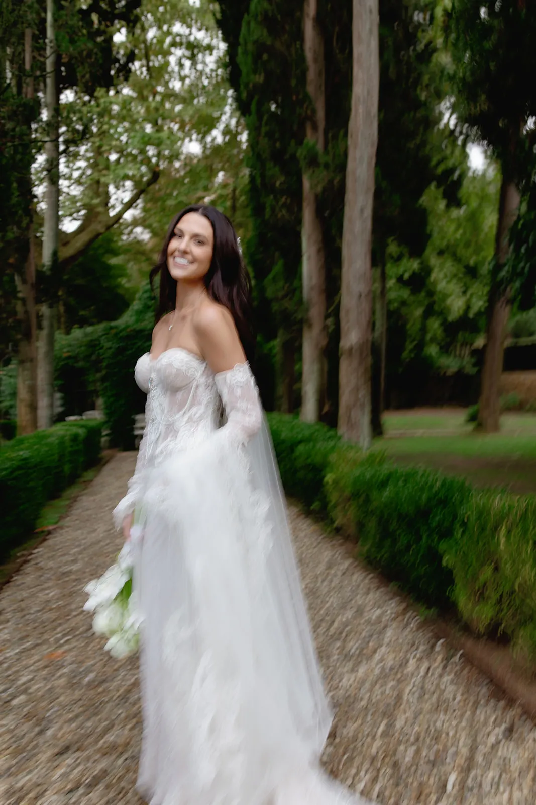 A smiling bride with long dark hair wears a strapless, lace wedding gown with sheer sleeves. She holds a white bouquet and stands on a gravel path bordered by neatly trimmed hedges and tall trees in a lush, green garden. Tuscan Wedding Photographer