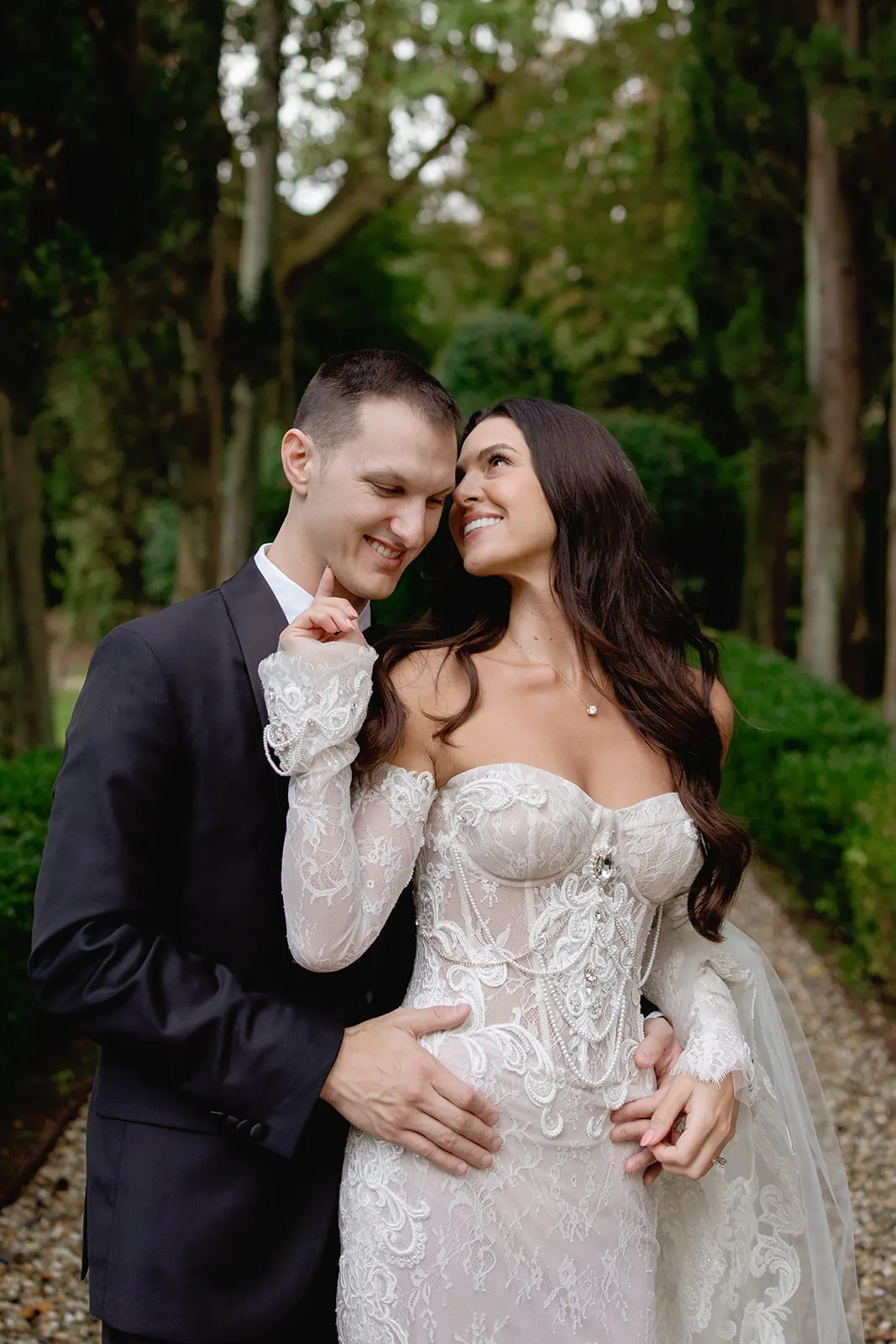 A bride in a long-sleeved lace wedding gown smiles at her groom, who wears a black suit and leans his forehead to hers. They embrace in a lush garden with tall trees and hedges, both looking joyful and affectionate. Tuscan Wedding Photographer