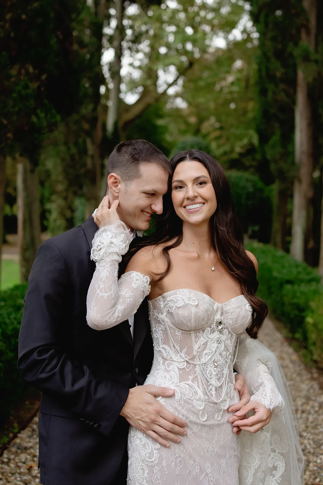A smiling bride in a lace off-shoulder wedding dress stands beside a groom in a dark suit. The groom embraces her waist and nuzzles her cheek. They are outside on a tree-lined path with greenery and sunlight in the background. Tuscan Wedding Photographer