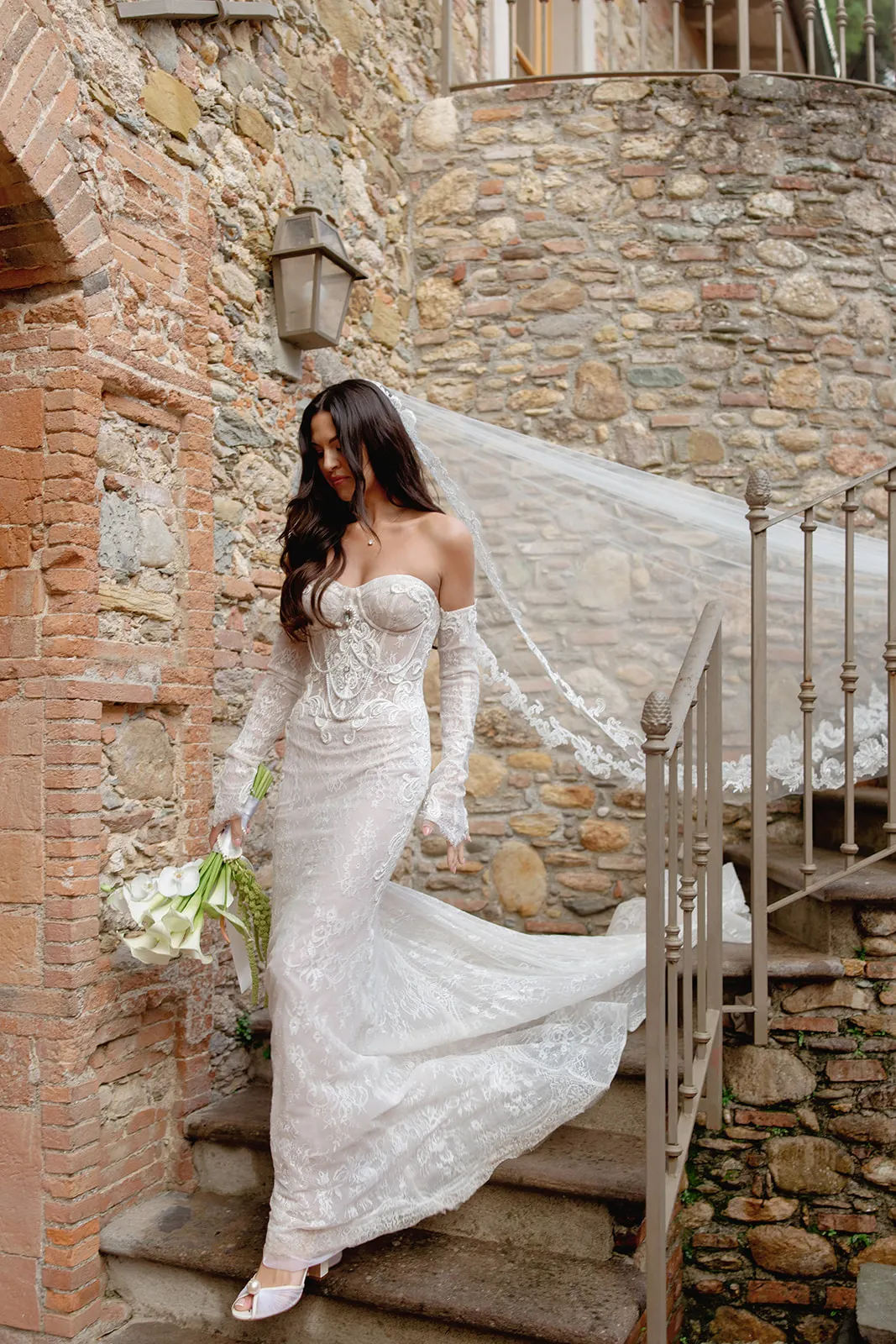 A bride in a fitted, off-the-shoulder lace wedding dress and long veil descends stone steps. She holds a bouquet of white flowers and has long dark hair, surrounded by rustic stone and brick walls in an outdoor setting. Tuscan Wedding Photographer
