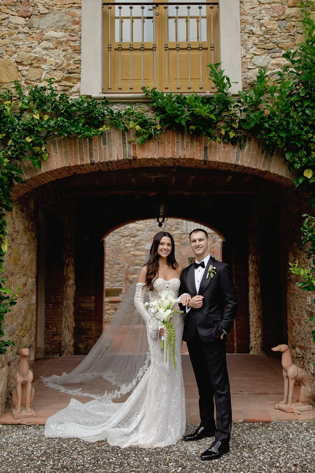 A bride in a white lace gown and veil stands beside a groom in a black tuxedo. They pose under a brick archway of a rustic stone building, holding hands and flowers. Two decorative dog statues are at their sides and greenery frames the scene. Tuscan Wedding Photographer