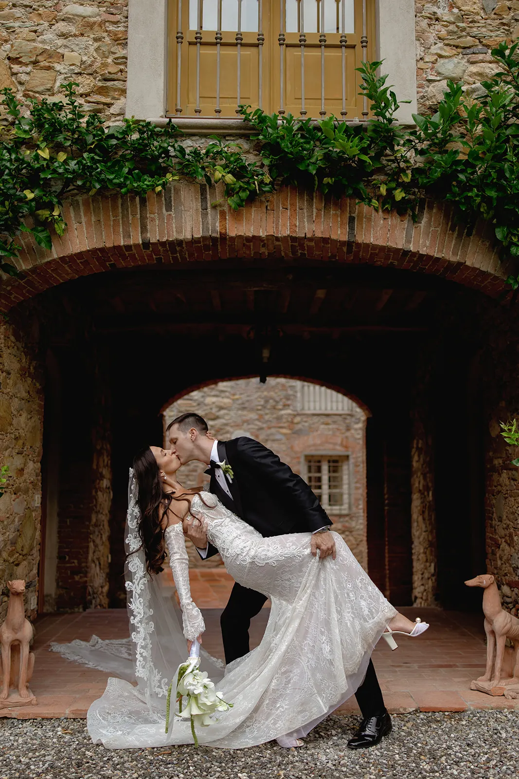 A groom in a black suit dips and kisses a bride in a white lace gown and veil. They are under a brick archway with green vines, flanked by stone walls and two dog statues. The bride holds a white bouquet as they share a joyful, intimate moment. Tuscan Wedding Photographer