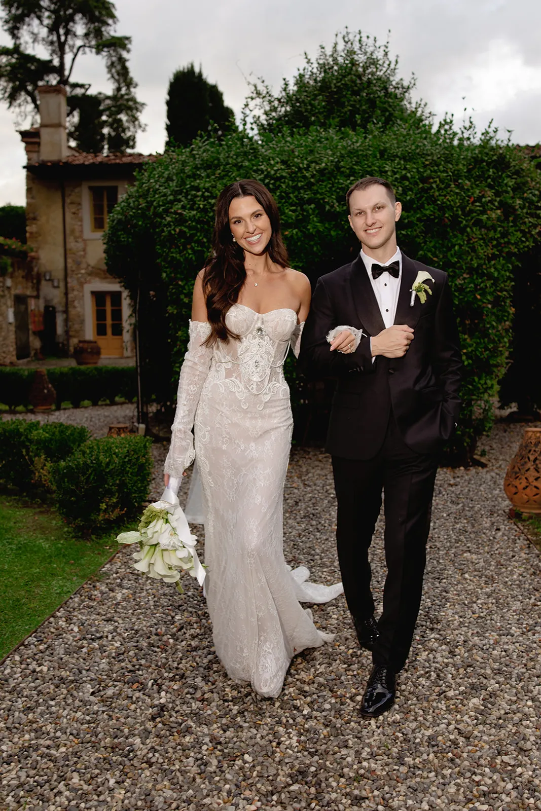 A bride in a fitted, off-shoulder lace gown and a groom in a black tuxedo walk arm-in-arm on a gravel path, smiling. The bride holds white flowers; lush greenery and a rustic building are in the background, suggesting an outdoor wedding setting. Tuscan Wedding Photographer