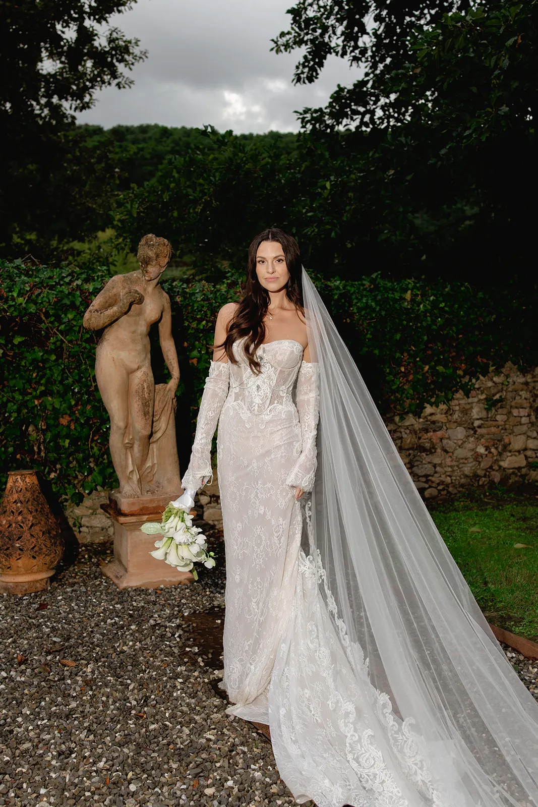 A bride in a long, lace off-the-shoulder wedding gown and veil stands on a gravel path, holding white flowers. She is next to a classical nude statue, with green foliage and a stone wall in the background under a cloudy sky. Tuscan Wedding Photographer