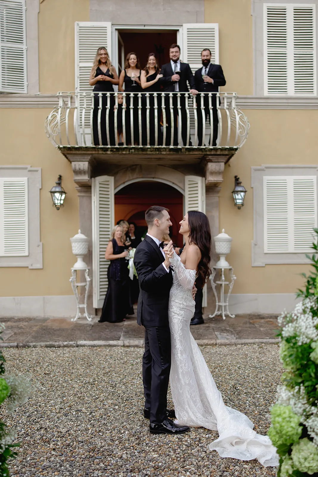 A bride and groom, both in formal attire, share a joyful dance outdoors in front of a beige building with white shutters. Friends and family watch from a balcony above and a doorway behind, all dressed elegantly, celebrating the couple’s wedding. Tuscan Wedding Photographer