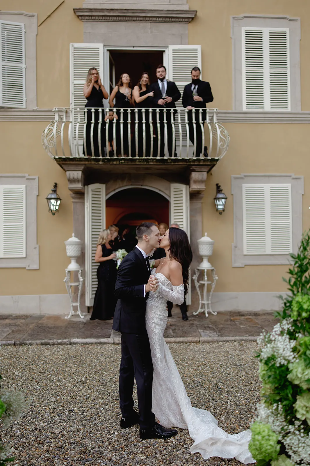 A bride and groom dance outdoors in front of a beige building with white shutters. Five well-dressed guests watch from a balcony above, while two more stand below. Greenery frames the scene, and the couple appears joyful and close. Tuscan Wedding Photographer