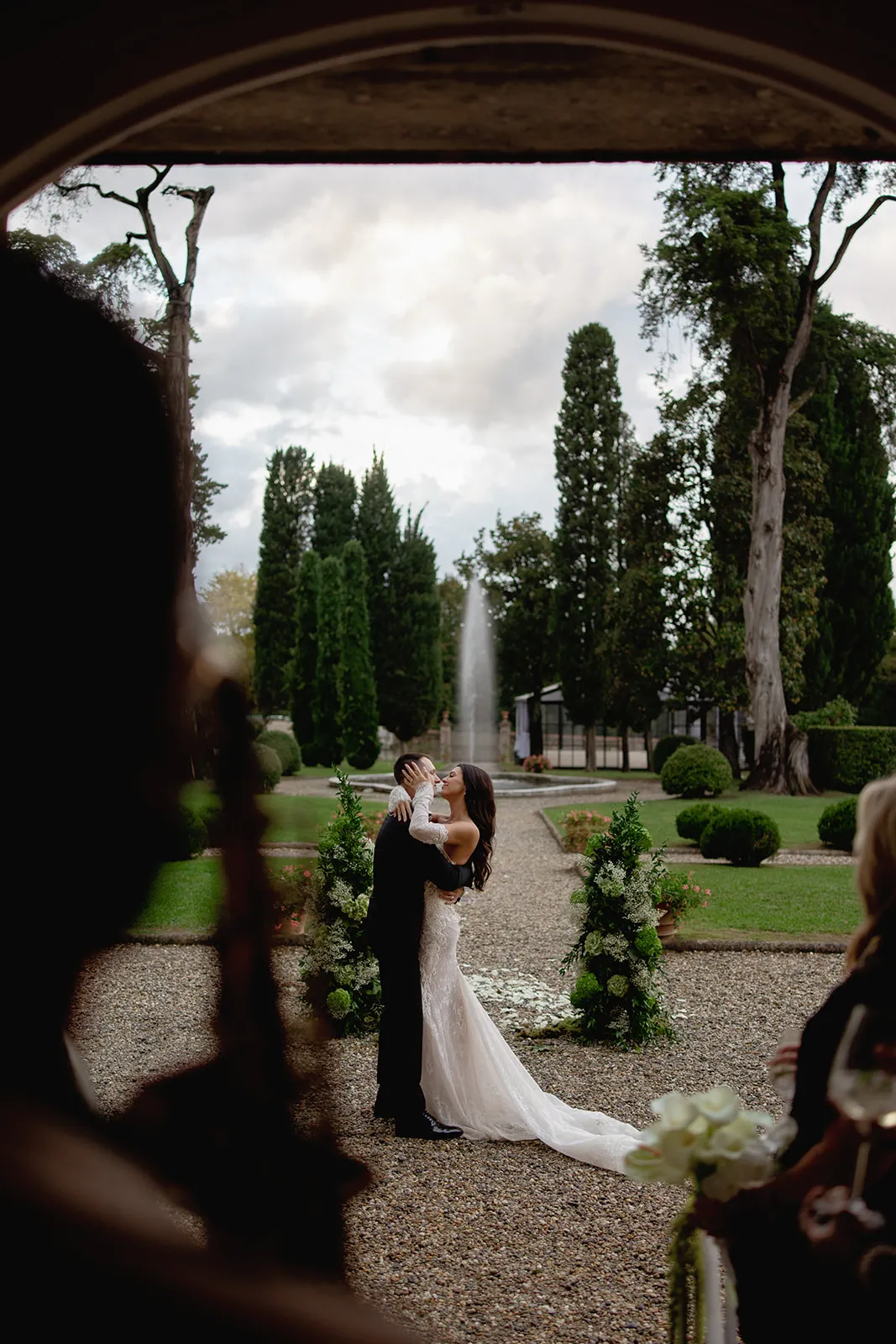 A bride and groom kiss outdoors during their wedding ceremony, framed by greenery and a tall fountain in the background, with guests watching in the foreground. The scene is elegant and romantic, set in a manicured garden with tall trees. Tuscan Wedding Photographer