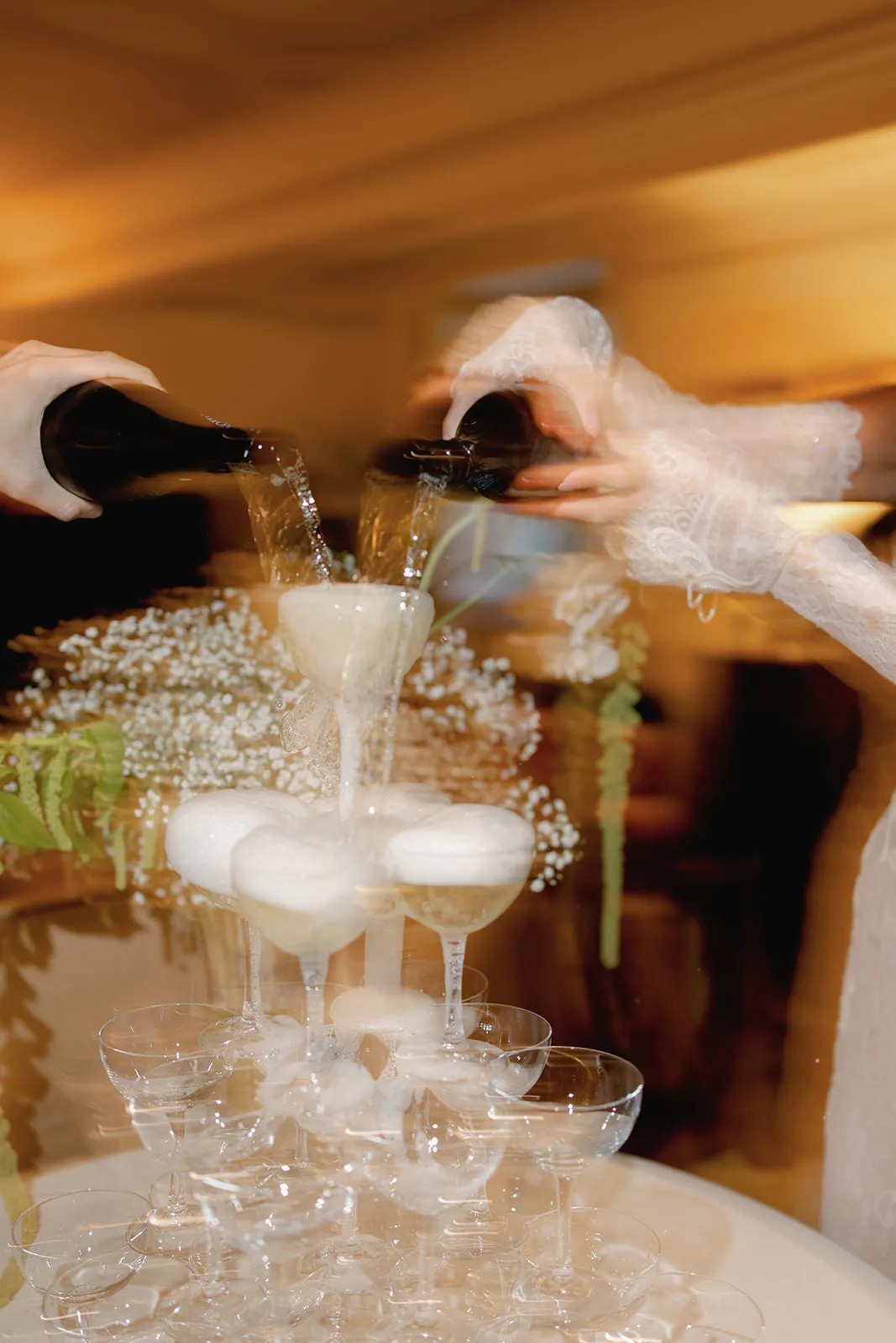 Two people pour champagne from dark bottles into the top glasses of a tiered champagne tower. Bubbles overflow as the champagne cascades down. Soft focus floral decorations are visible in the background. The scene looks elegant and celebratory. Tuscan Wedding Photographer