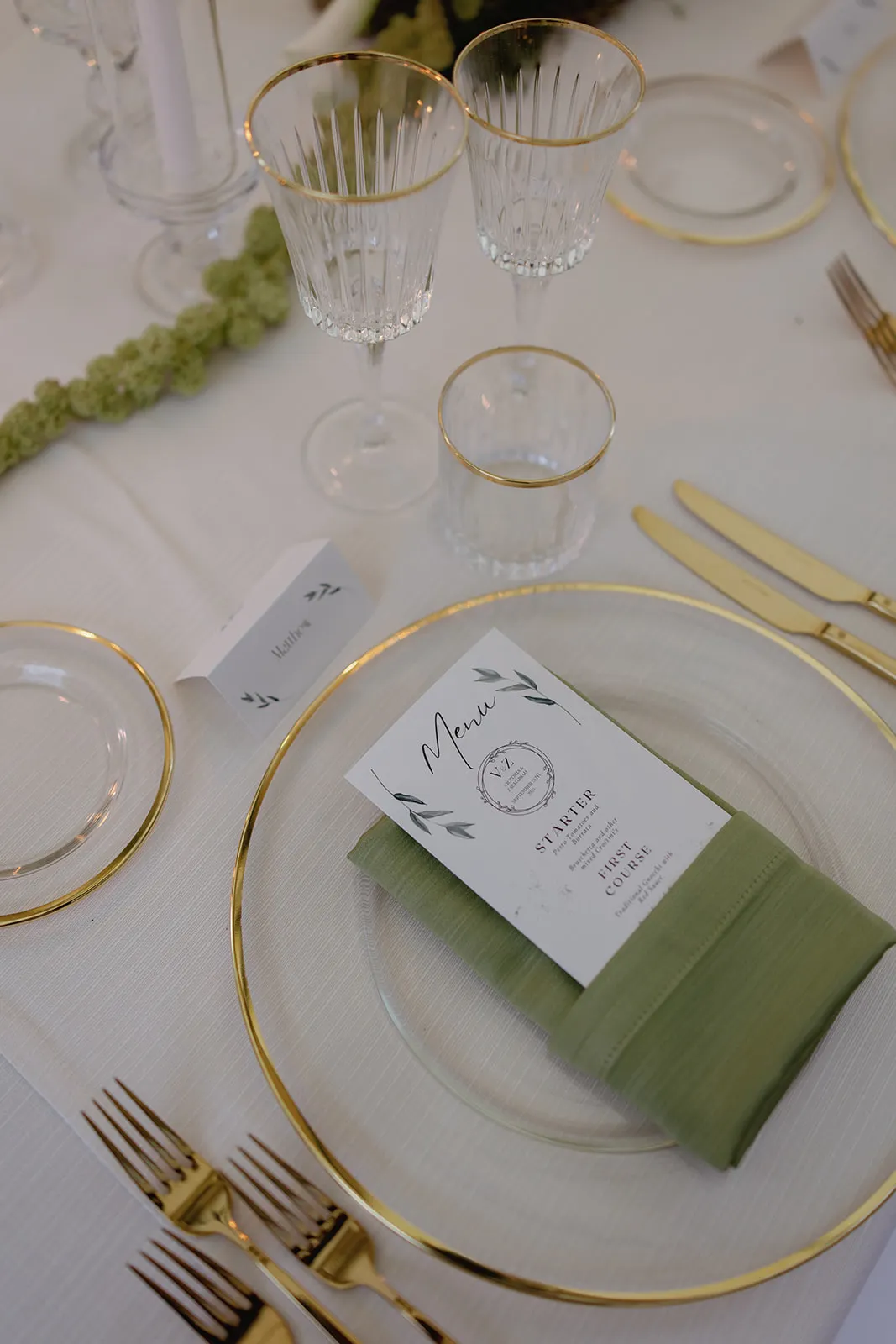 Elegant table setting with a clear plate rimmed in gold, a green napkin, and a menu card on top. Gold flatware and rimmed glasses surround the plate. A place card and green garland decorate the white tablecloth background. Tuscan Wedding Photographer
