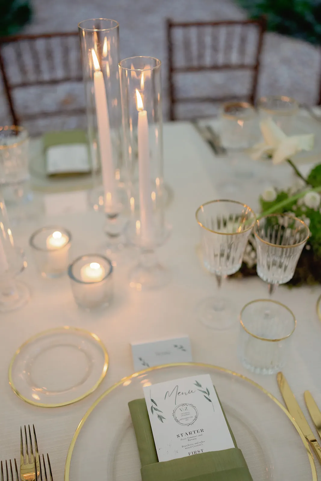Elegant table setting with clear plates edged in gold, gold utensils, crystal wine glasses, tall glass candle holders with lit white candles, and small votive candles. A green napkin and a printed menu rest on the plate; greenery decorates the table. Tuscan Wedding Photographer