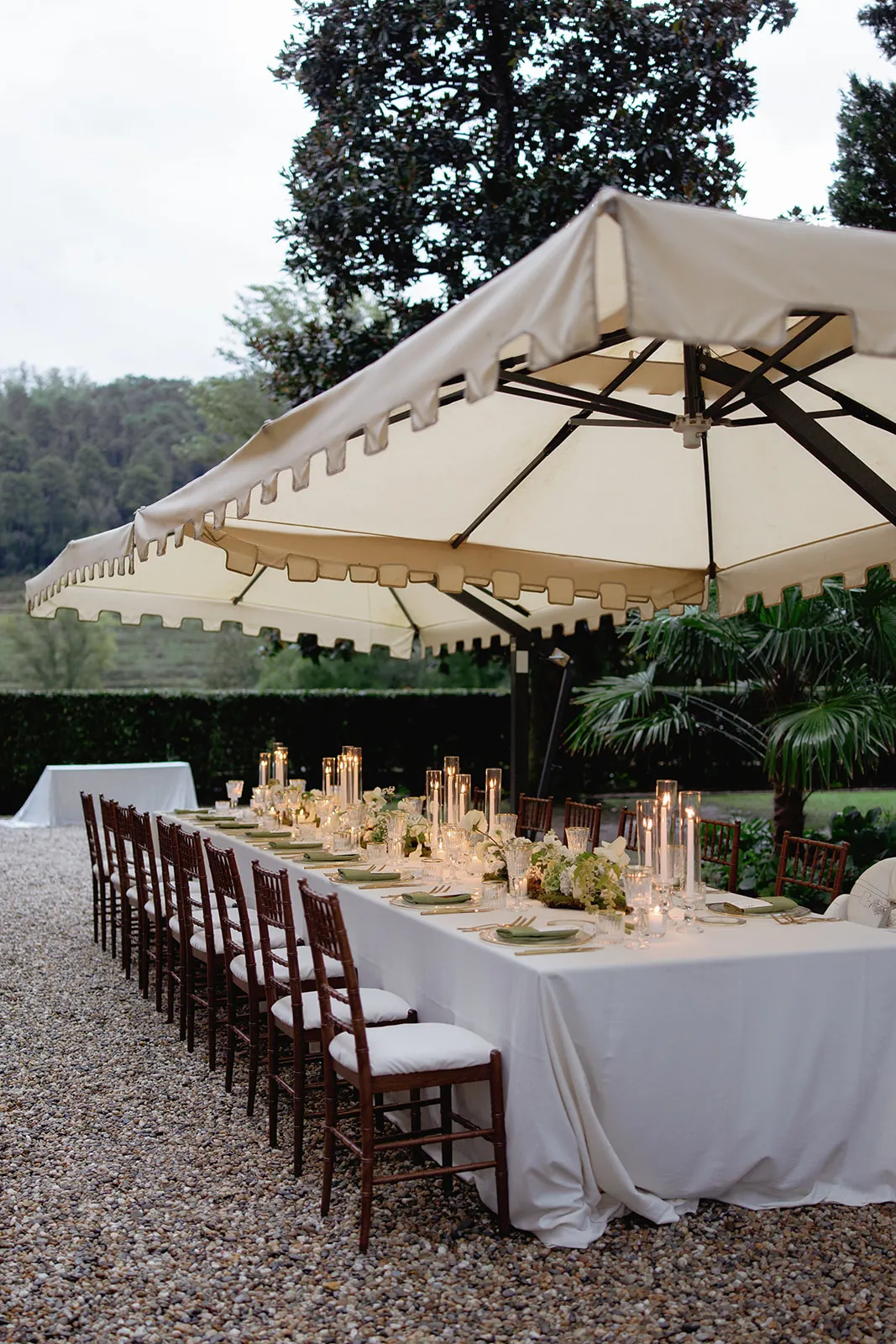 An elegant outdoor dining setup features two long tables with white tablecloths, brown wooden chairs, floral centerpieces, and lit candles under a large cream canopy. The scene is surrounded by greenery and trees, creating a serene garden atmosphere. Tuscan Wedding Photographer