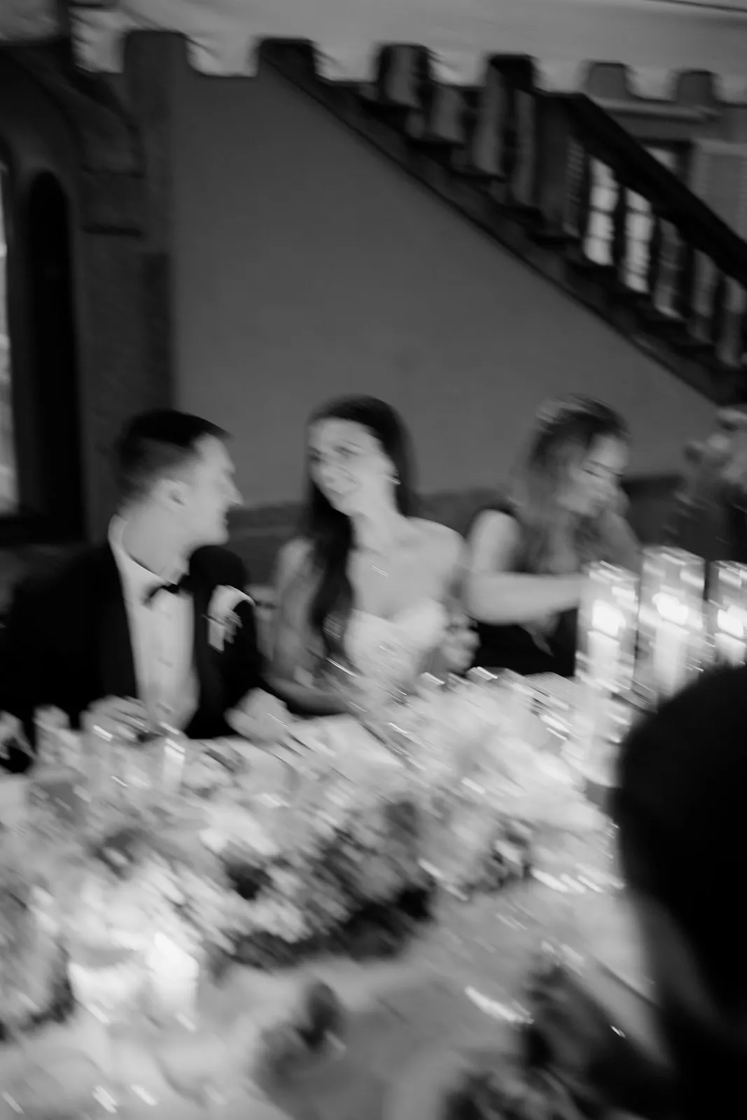 A black and white, slightly blurred photo of a formal dinner. A man in a suit and bow tie sits beside a woman in a strapless dress. They look at each other and smile. A third person sits next to them. Candles and flowers decorate the table. Tuscan Wedding Photographer