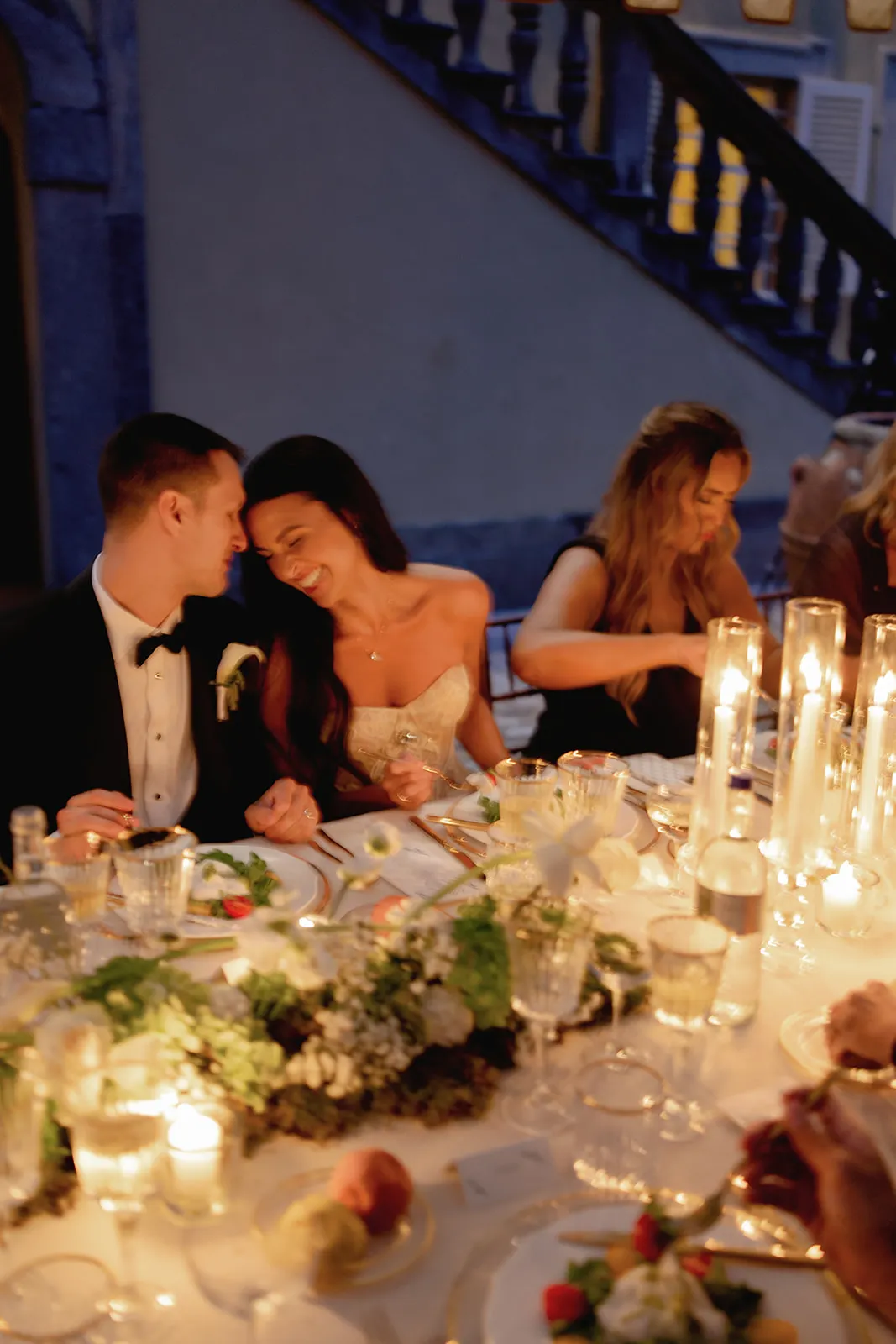A bride and groom sit close together, smiling affectionately, at a candlelit dinner table decorated with flowers and greenery. Guests around them enjoy their meal in a warm, elegant setting with soft lighting and tall glass candles. Tuscan Wedding Photographer