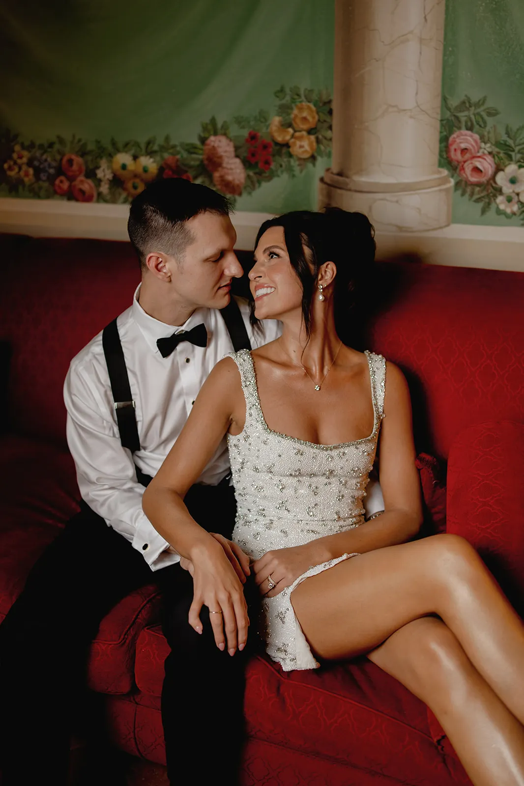 A couple sits on a red couch, gazing at each other and smiling. The man wears a white shirt, black suspenders, and a bow tie. The woman wears a short, sleeveless white dress with beaded details. Behind them is a painted floral mural and a white column. Tuscan Wedding Photographer