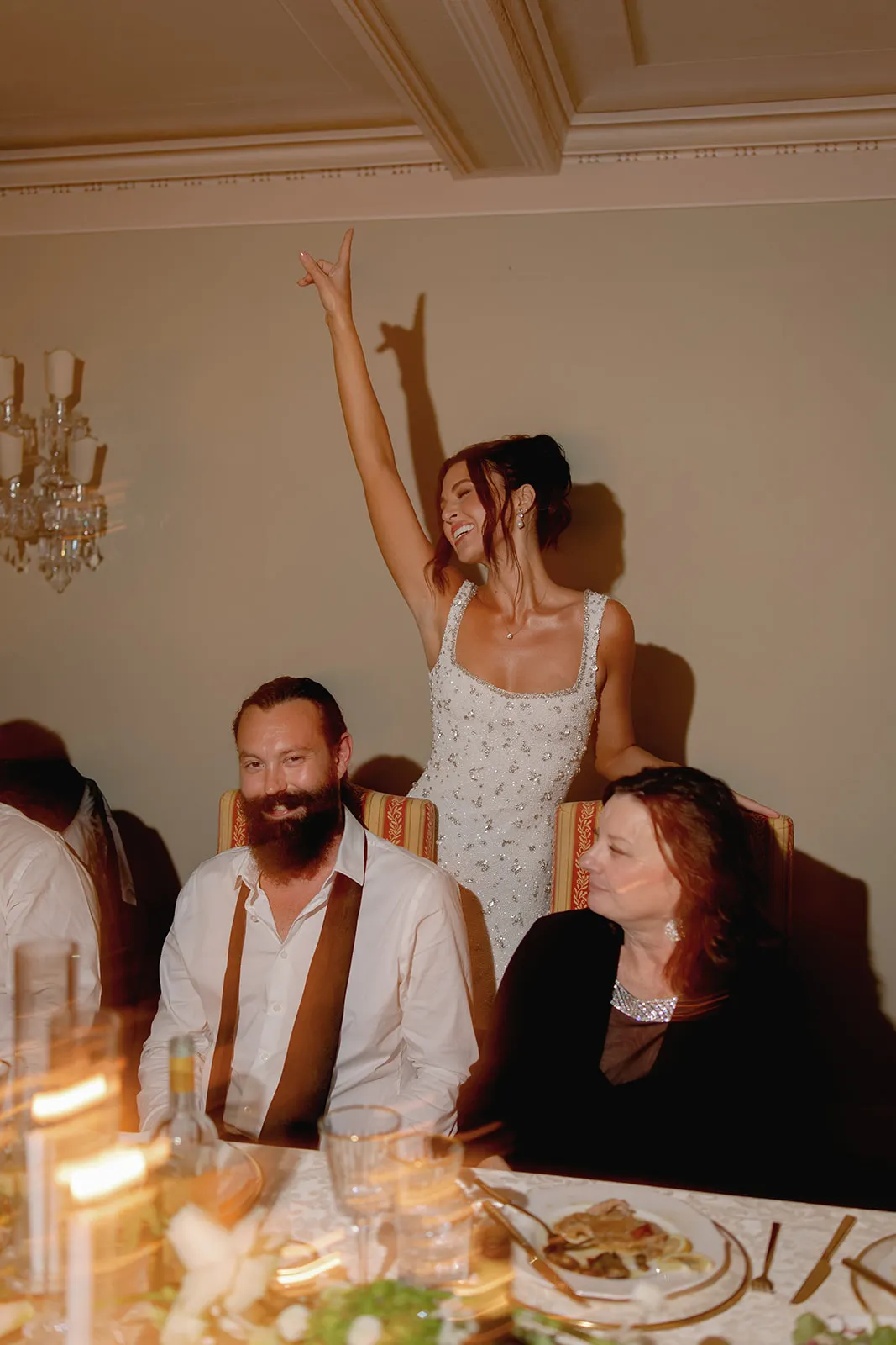 A smiling woman in a white, beaded dress stands behind a seated man and woman at a dining table. She raises one arm enthusiastically, fingers forming a rock-and-roll gesture. The table is set with plates, glasses, a bottle, and blurred candles in the foreground. Tuscan Wedding Photographer