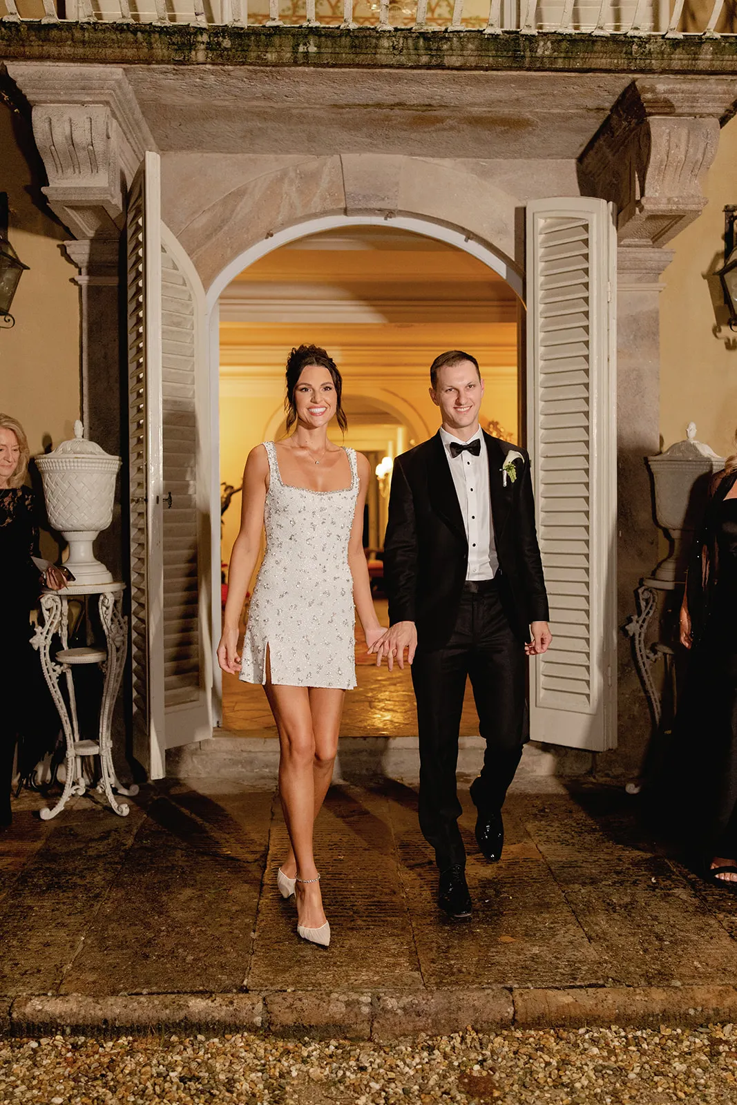 A smiling couple holding hands walks out of open double doors at night. The woman wears a short white dress and heels, and the man wears a black tuxedo with a white shirt and bow tie. Guests in dark attire stand nearby, watching them. The setting looks elegant and festive. Tuscan Wedding Photographer