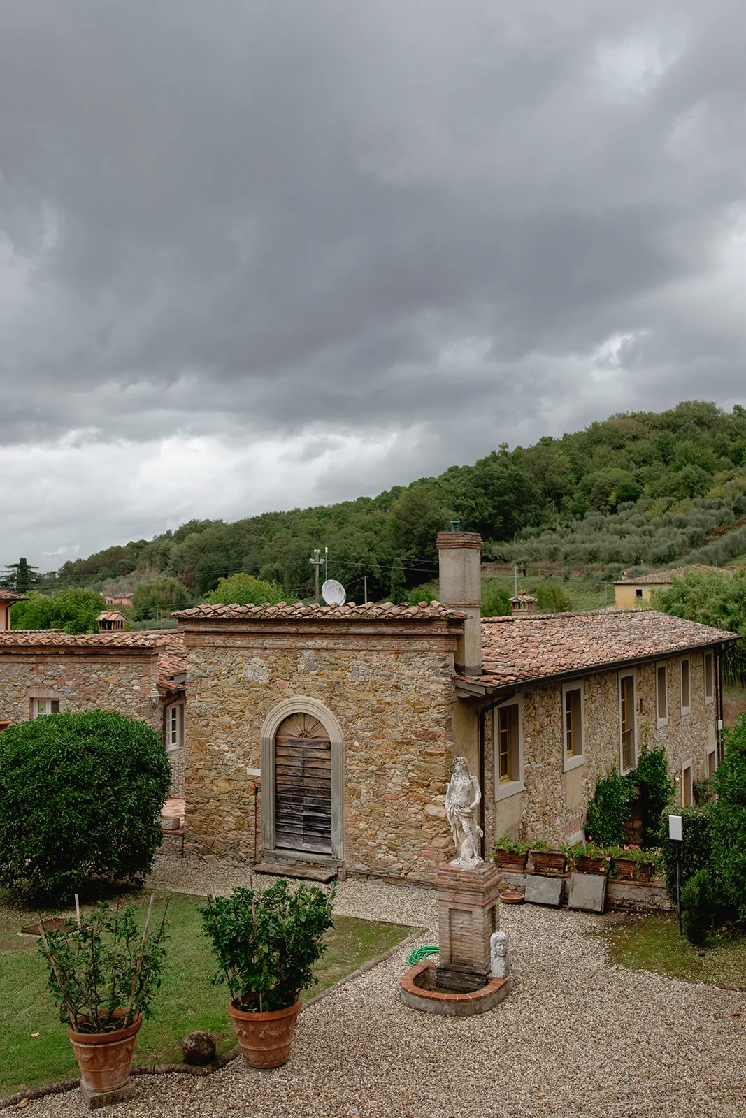A rustic stone building with a red-tiled roof stands under a cloudy sky. There is a statue in front, potted plants, and green hedges. Behind the building, a lush, tree-covered hill rises, giving the scene a peaceful, countryside feel. Tuscan Wedding Photographer