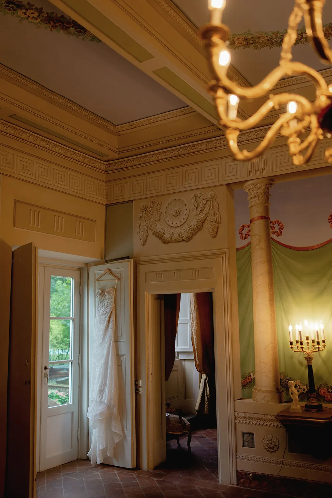 A white lace wedding dress hangs on a door in an elegant, ornately decorated room with high ceilings, intricate molding, and a lit chandelier. Soft daylight enters through the open window, highlighting lush greenery outside. Tuscan Wedding Photographer