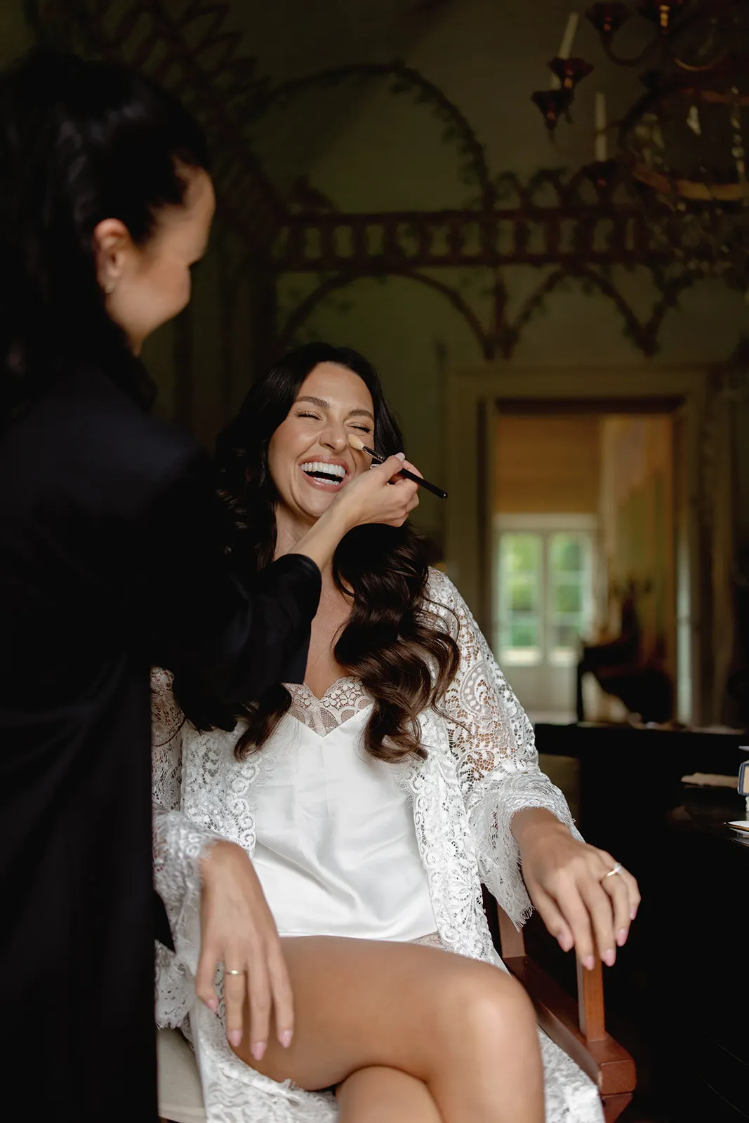 A woman in a white lace robe and slip sits smiling with eyes closed as another person applies makeup to her face. The room is dimly lit with ornate walls and a window in the background, creating a cozy and elegant atmosphere. Tuscan Wedding Photographer