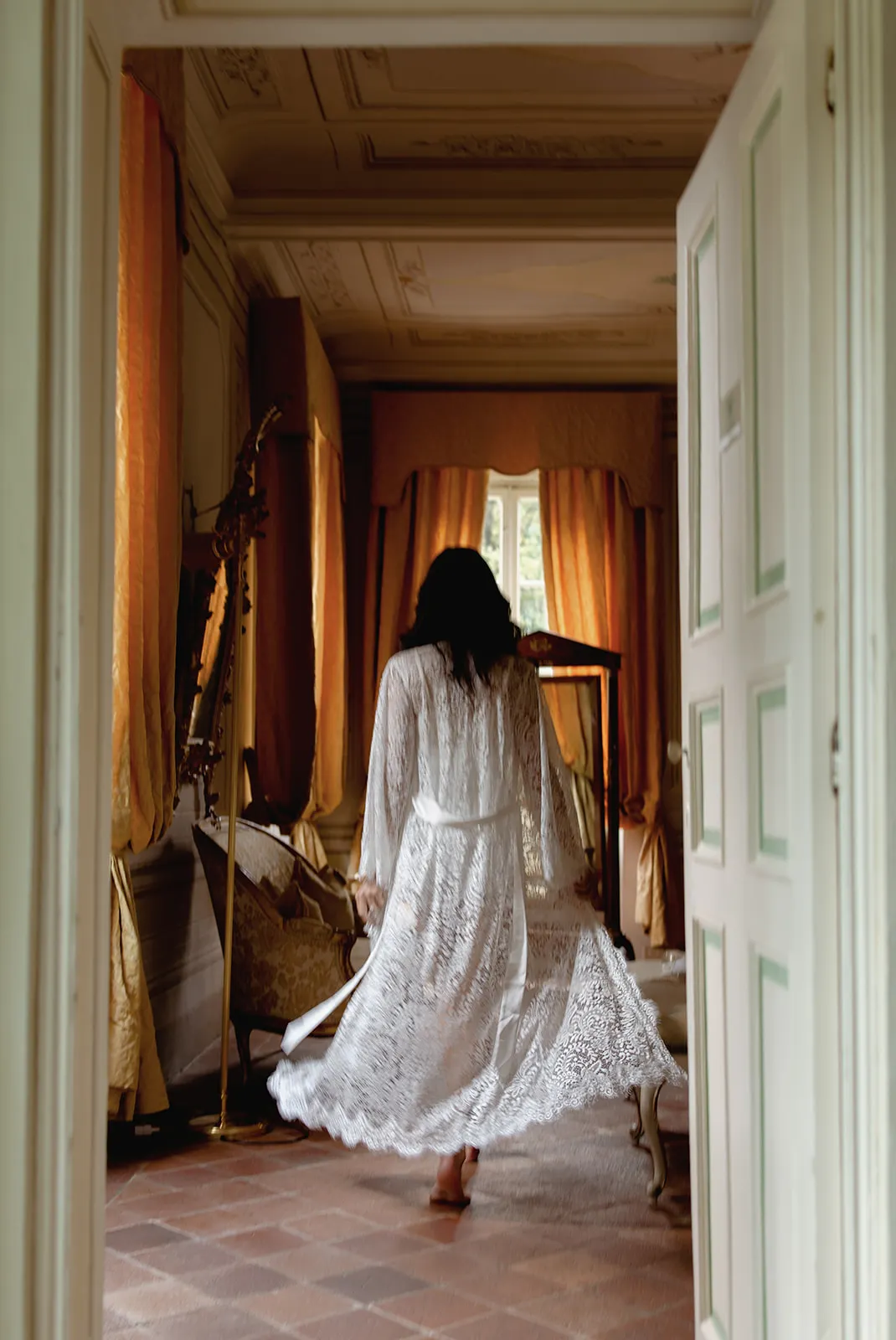 A person in a long, white lace robe walks barefoot through an elegant, vintage room with tall windows, gold curtains, and ornate furniture. They are seen from behind, swirling slightly, with natural light filtering in through the curtained windows. Tuscan Wedding Photographer