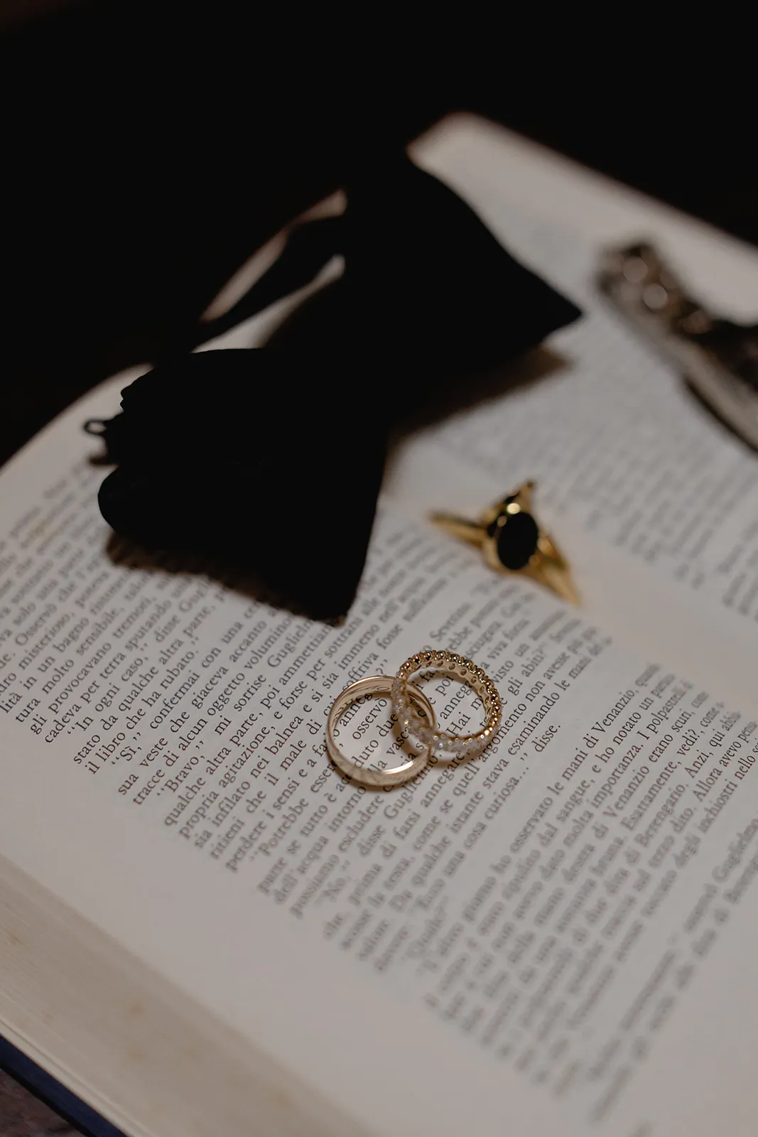 Two gold rings rest on the open pages of a book with text in a serif font. Nearby are a black bow tie and a gold ring with a dark stone, all arranged on the book, suggesting a formal or romantic occasion. Tuscan Wedding Photographer
