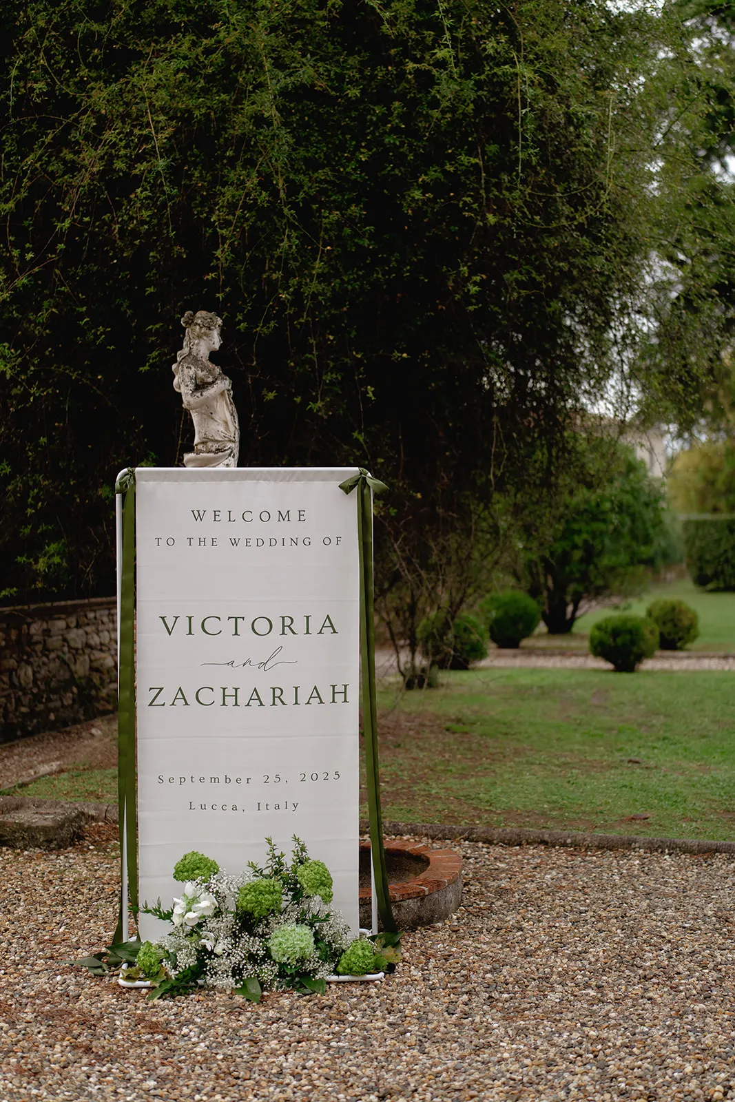 A white wedding welcome sign with green ribbon stands outdoors on gravel, surrounded by greenery and flowers. The sign reads: “Welcome to the wedding of Victoria and Zachariah, September 29, 2023, Lucca, Italy.” A small statue is on top of the sign. Tuscan Wedding Photographer