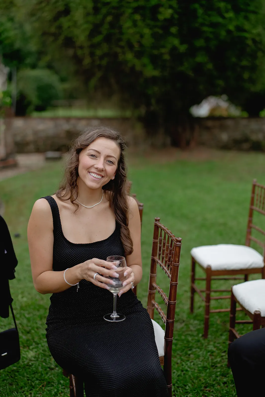 A woman in a sleeveless black dress sits on a wooden chair outdoors on grass, smiling at the camera while holding a wine glass. She has long brown hair and wears a necklace and bracelets. Empty chairs and greenery are visible in the background. Tuscan Wedding Photographer