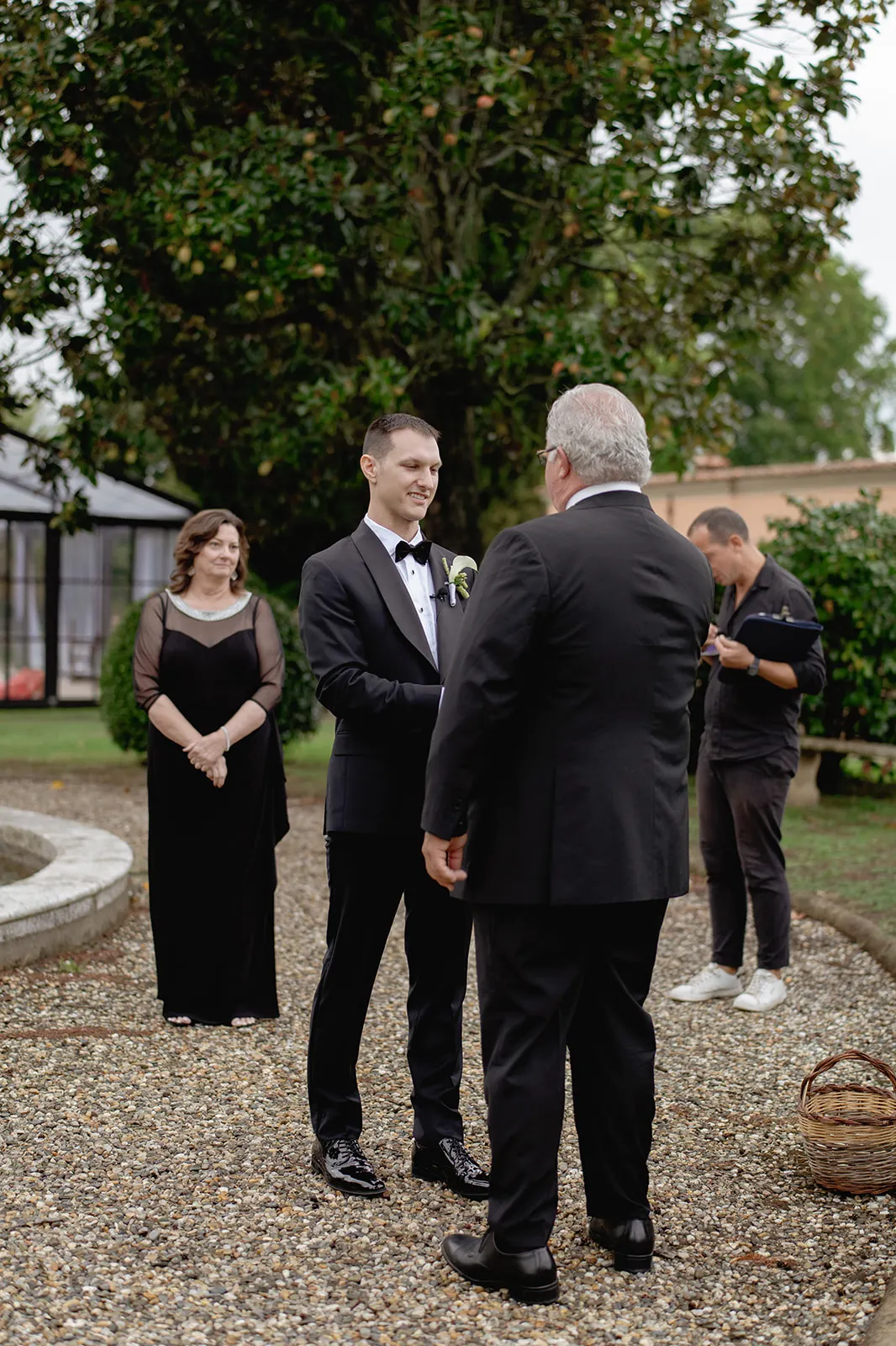 A groom in a black tuxedo smiles while holding hands with an older man in a black suit outdoors. A woman in a black dress watches and smiles nearby. Another man, casually dressed, stands in the background near greenery and a tree. Tuscan Wedding Photographer