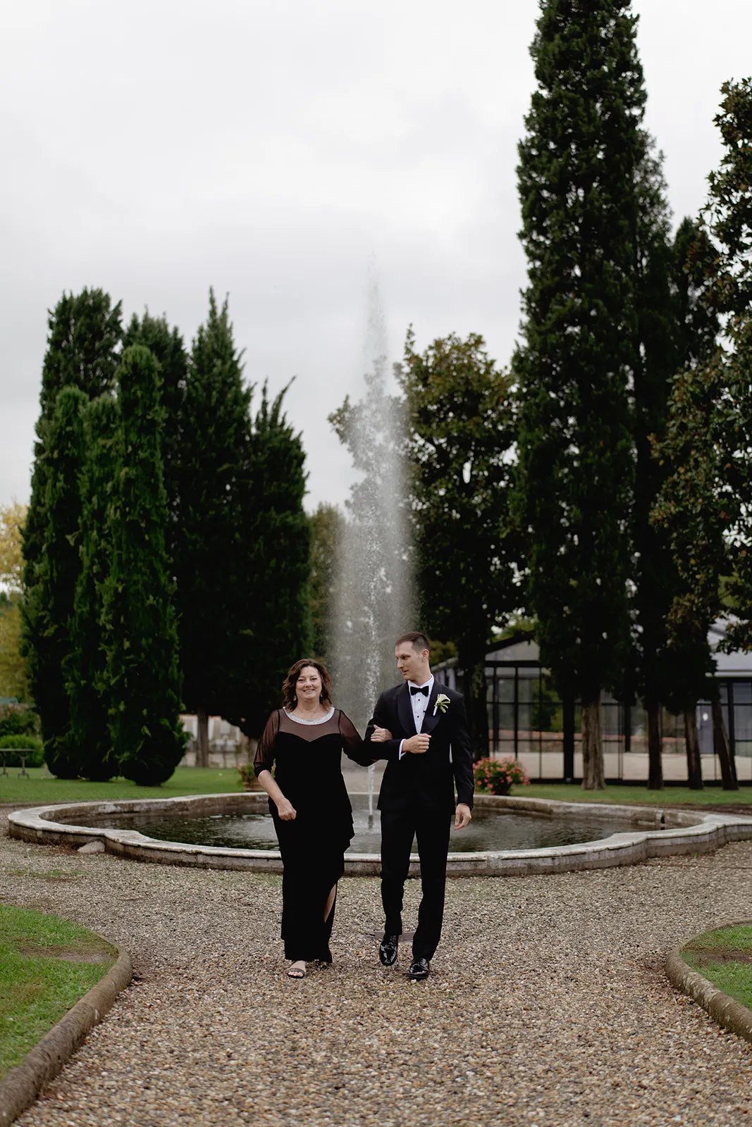A woman in a long black dress and a man in a black tuxedo walk arm-in-arm on a gravel path in front of a water fountain, surrounded by tall green trees and manicured gardens under an overcast sky. Tuscan Wedding Photographer